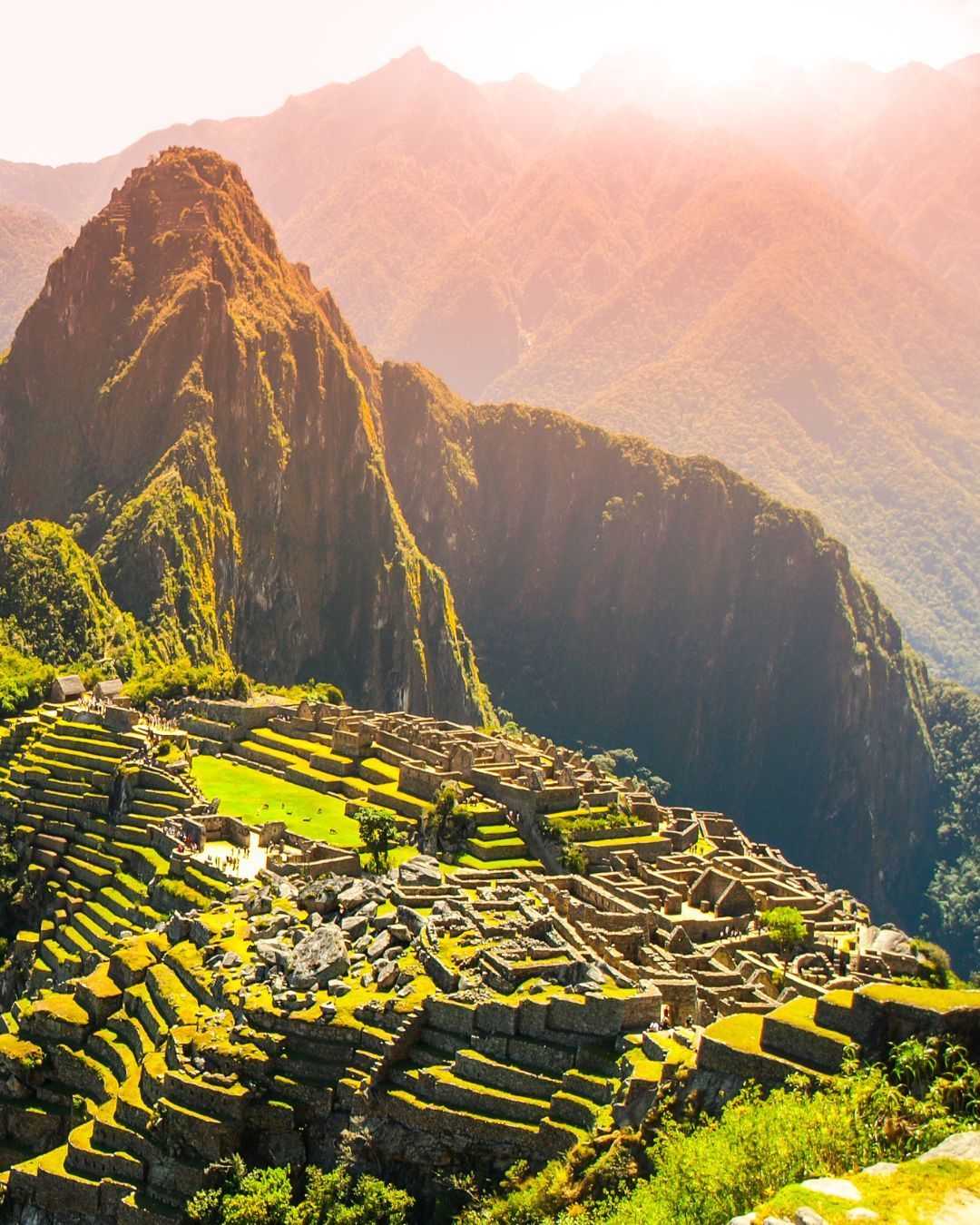 Sunlit Machu Picchu ruins on a mountainside with terraced fields and misty peaks in the background