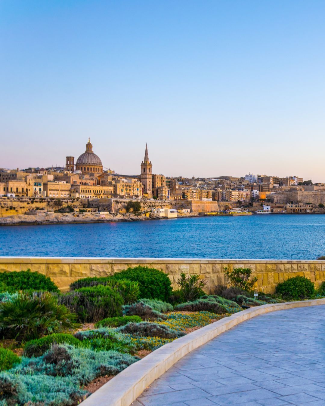Sunlit waterfront cityscape with a domed cathedral, blue water, and a landscaped promenade in the foreground