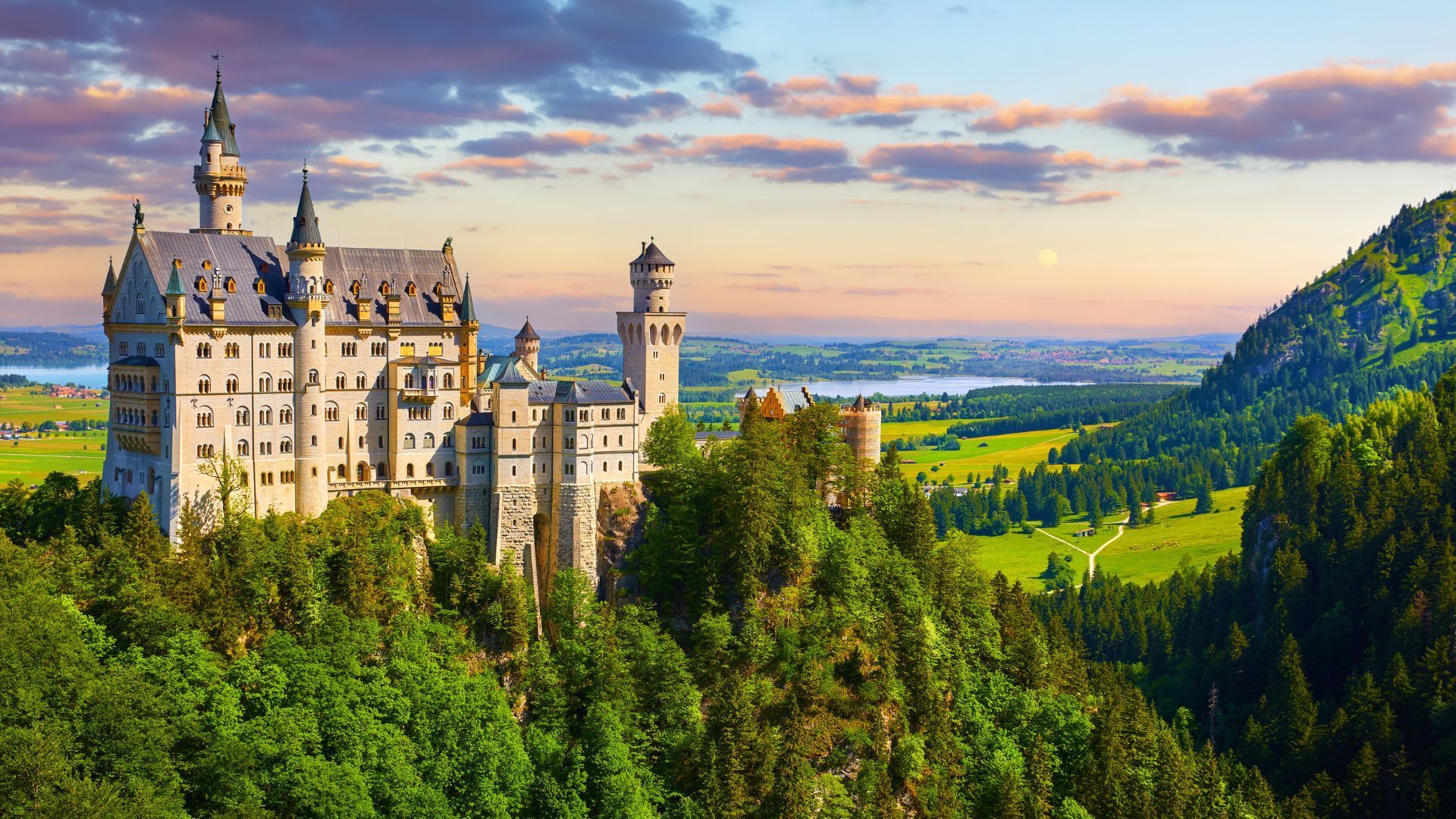 Neuschwanstein Castle perched on a hill, surrounded by green forest and overlooking a valley and lake, at sunset.