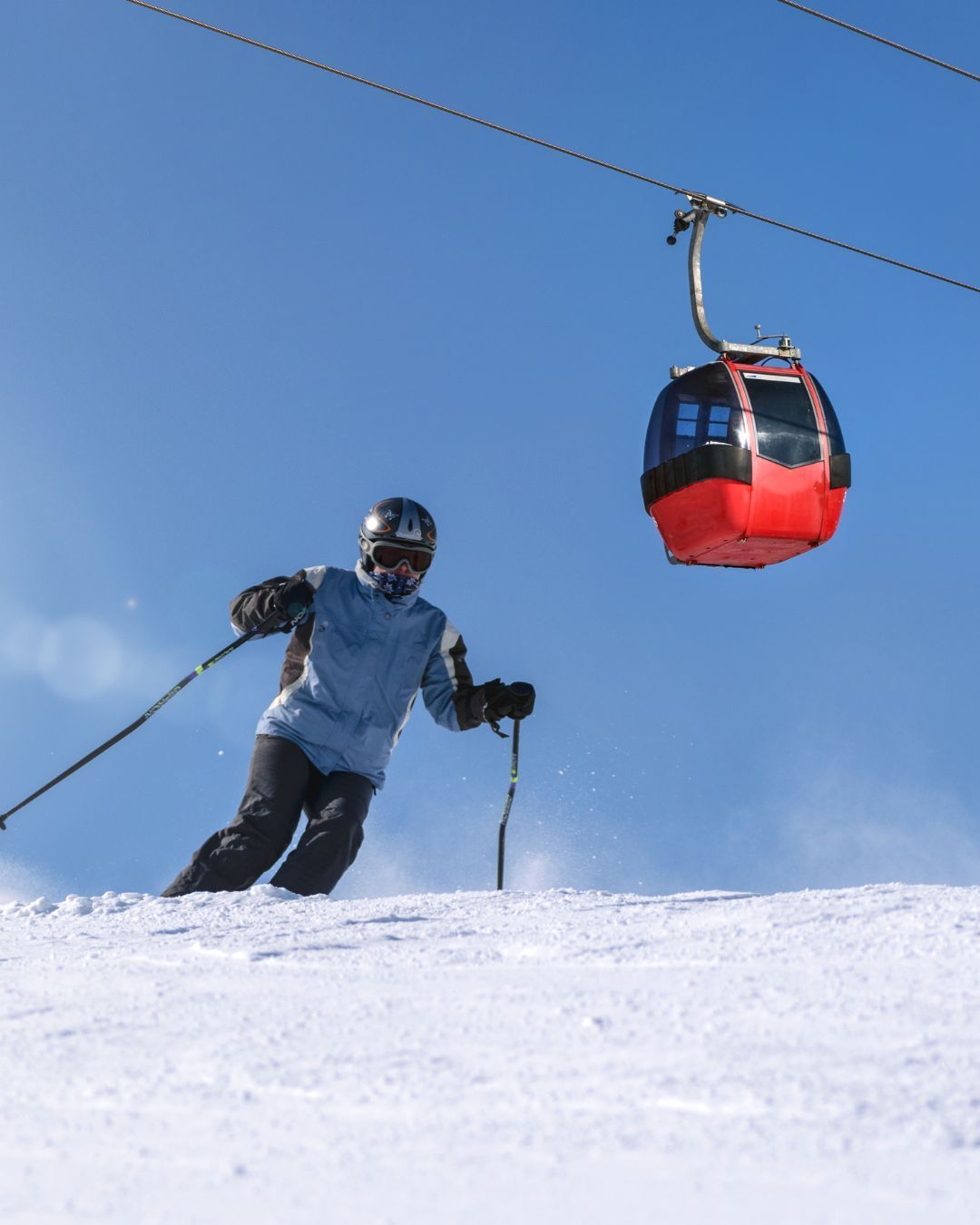 Skier in blue jacket carving snow under a red gondola on a clear mountain slope