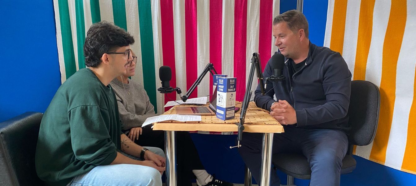 Two men are sitting at a table talking to each other in front of a microphone.