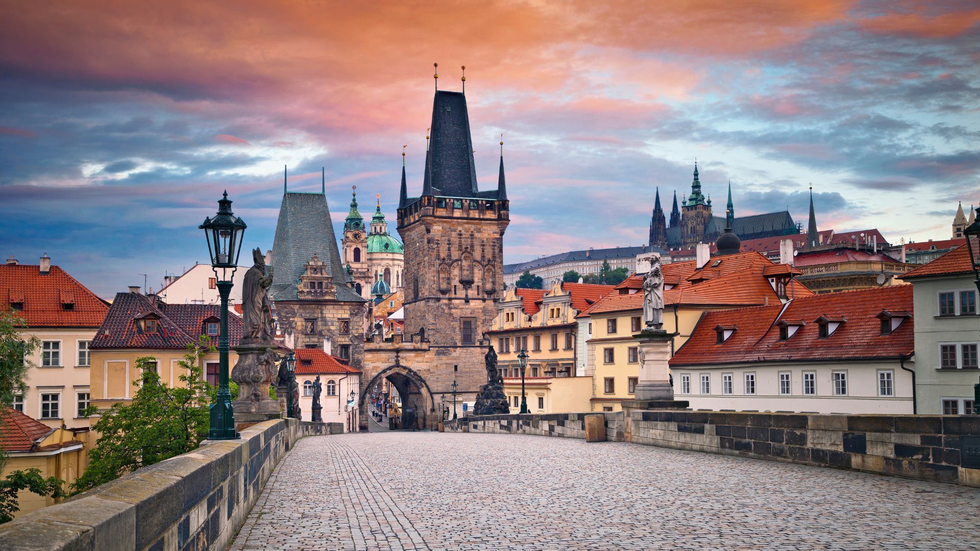 Cobblestone bridge leading to Prague's historic buildings, under a colorful sunset sky.