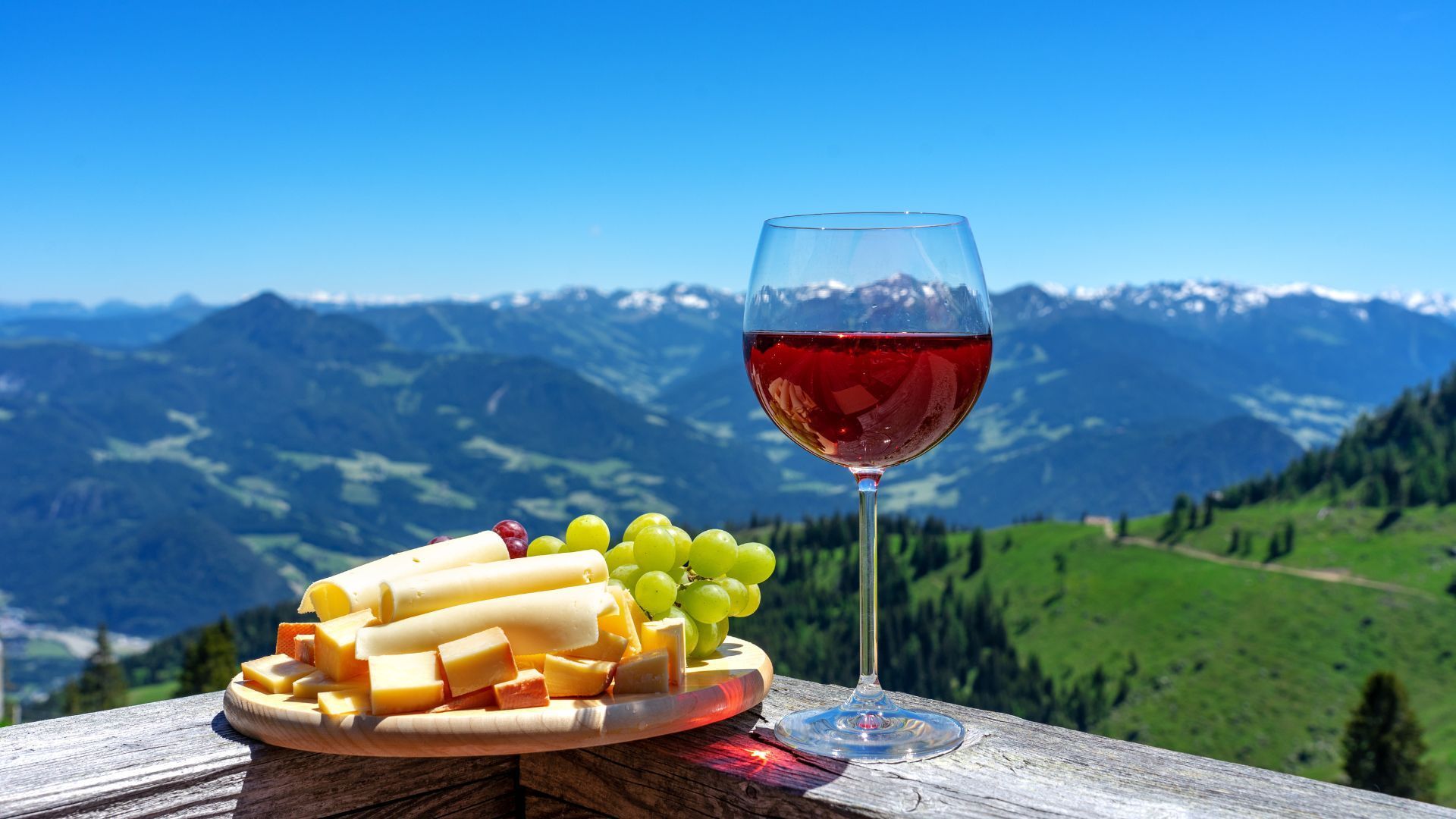 A glass of wine and a plate of cheese on a table with mountains in the background.