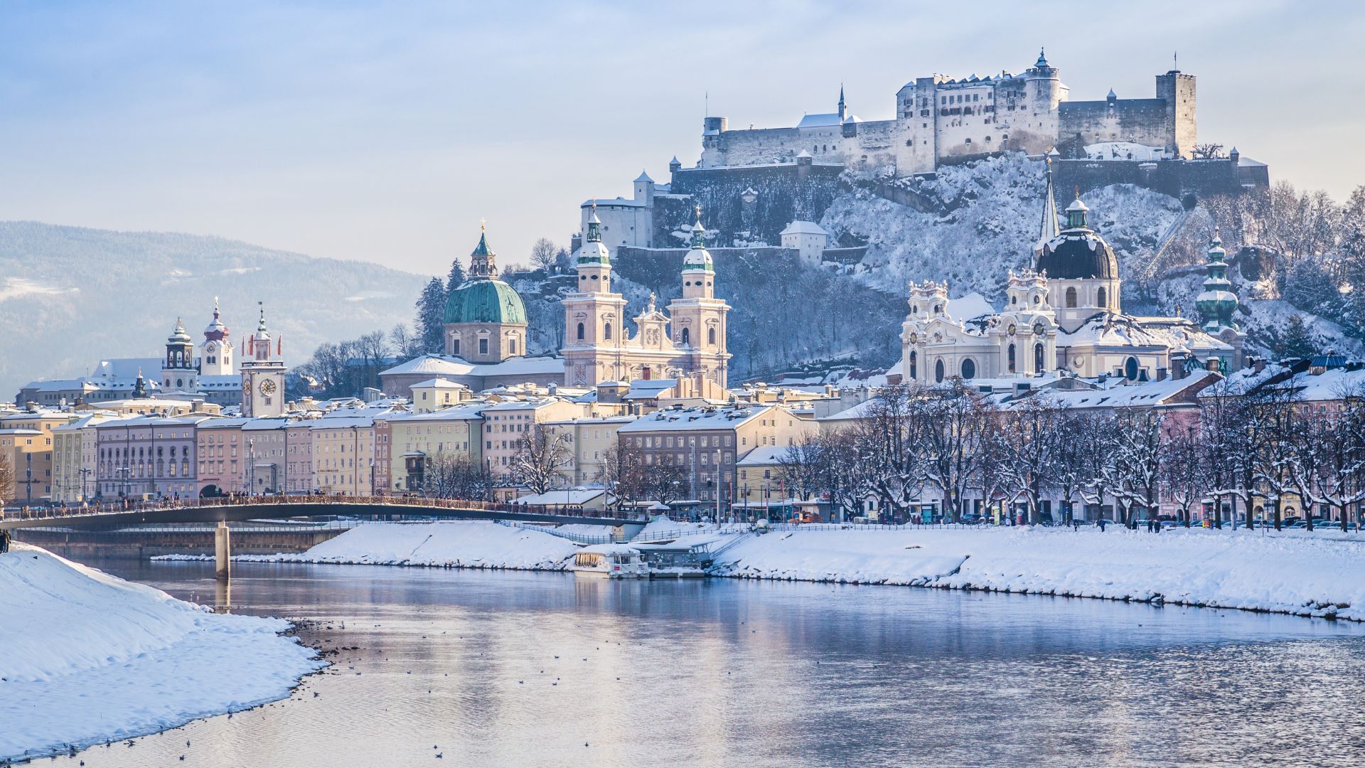 A snowy city with a castle in the background and a river in the foreground.