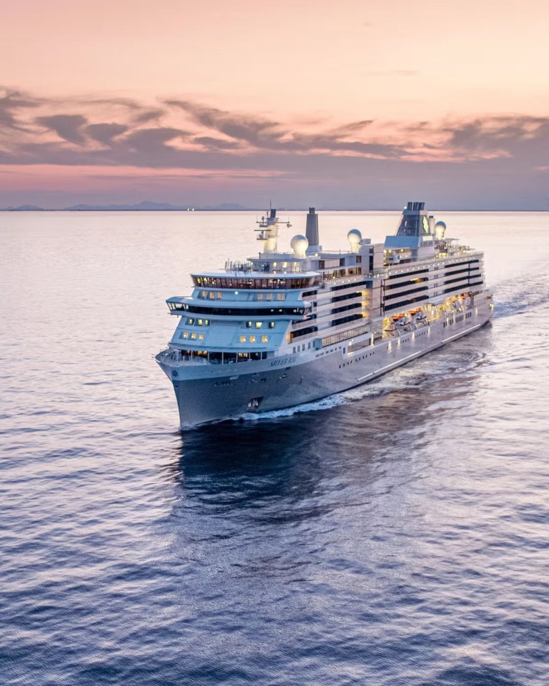 Large white cruise ship, Silversea Silver Ray, sailing on calm water at sunset, with a pink sky and distant horizon.