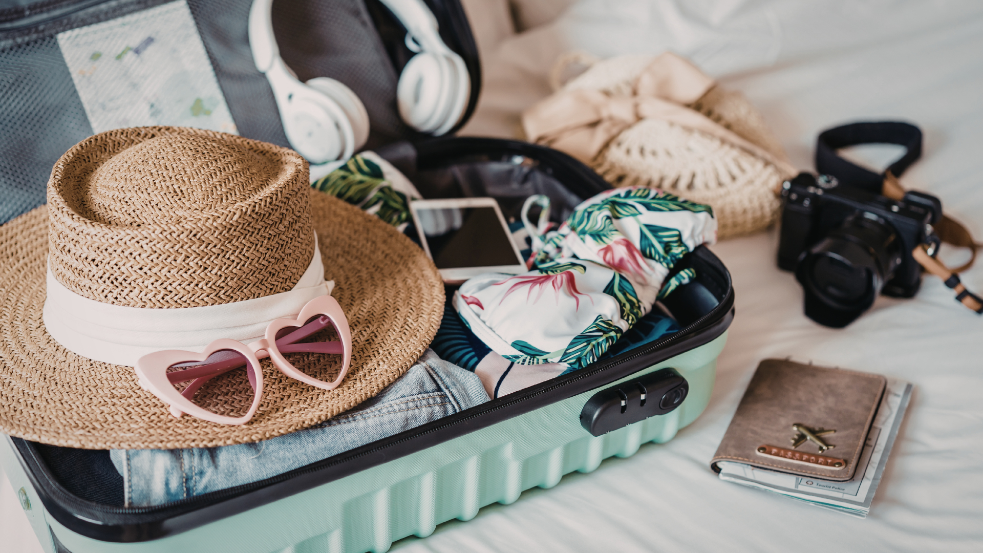 Open suitcase with hat, sunglasses, headphones, camera, phone, and clothing on a bed.
