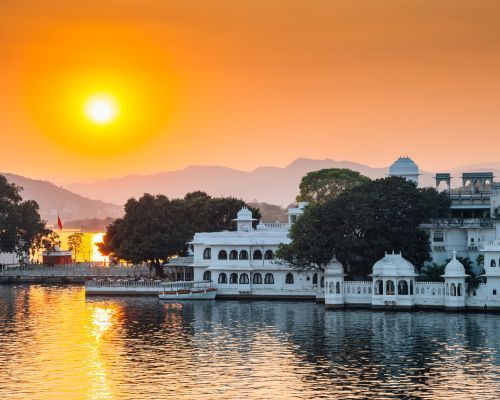 The sun is setting over a lake with buildings in the foreground and mountains in the background.
