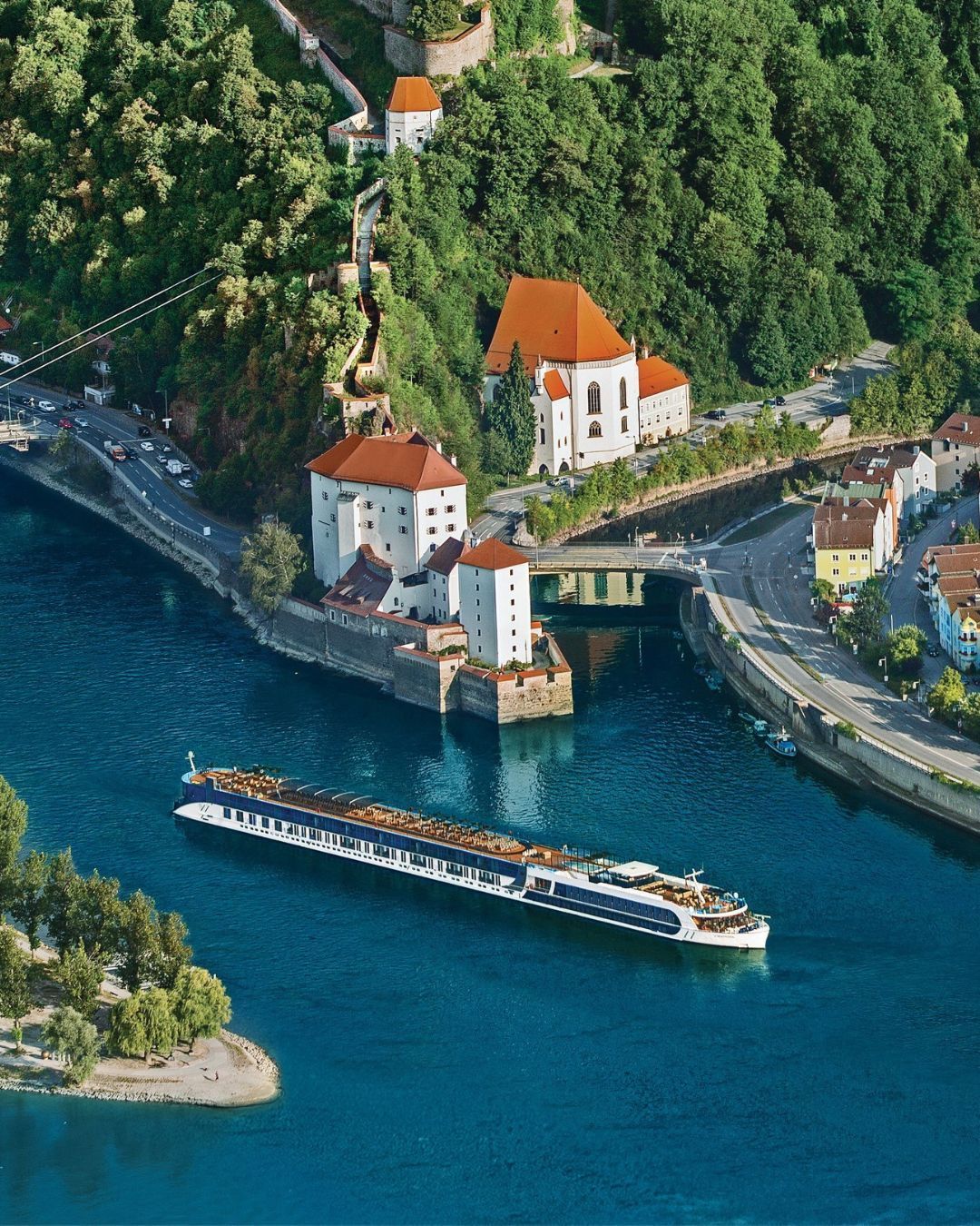 Aerial view of a riverboat passing a riverside village with red-roofed buildings and a forested cliff.