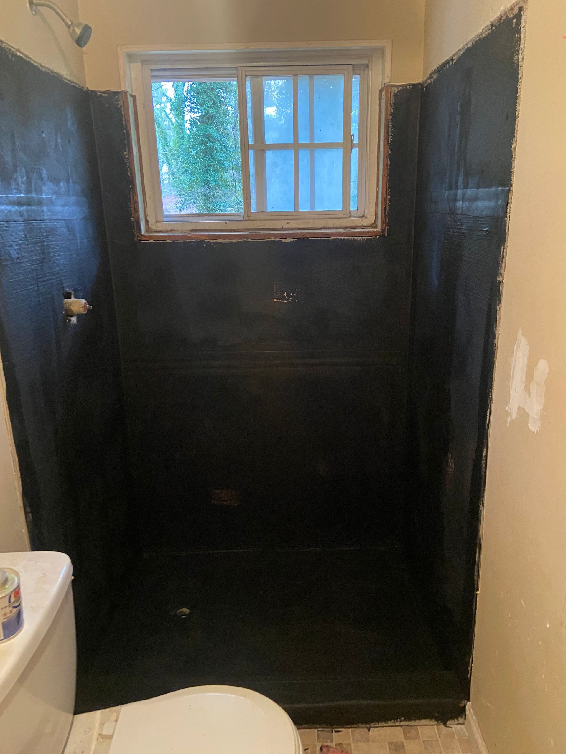 Black waterproofed shower stall, small window above. Toilet on the left, tan walls.