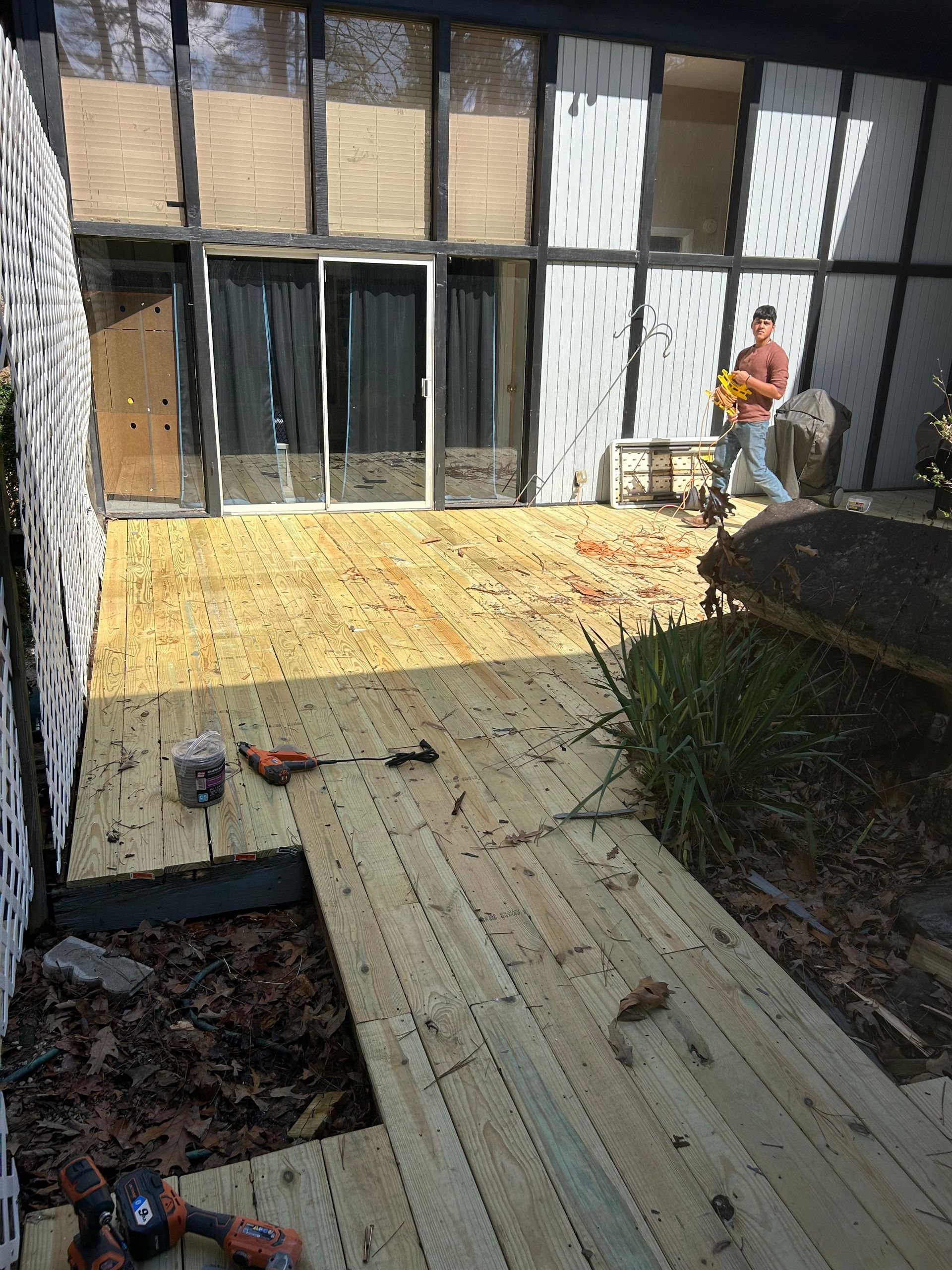 A wooden deck being built, with a person carrying tools in a modern setting with large windows.