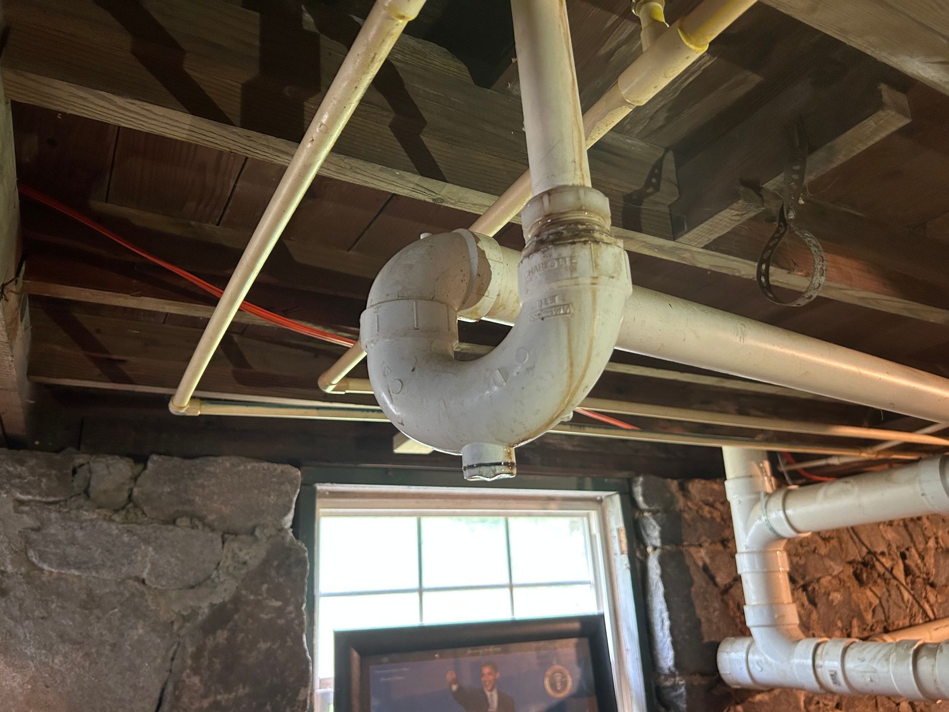 White pipe with a U-bend hangs from wooden ceiling near a small window.