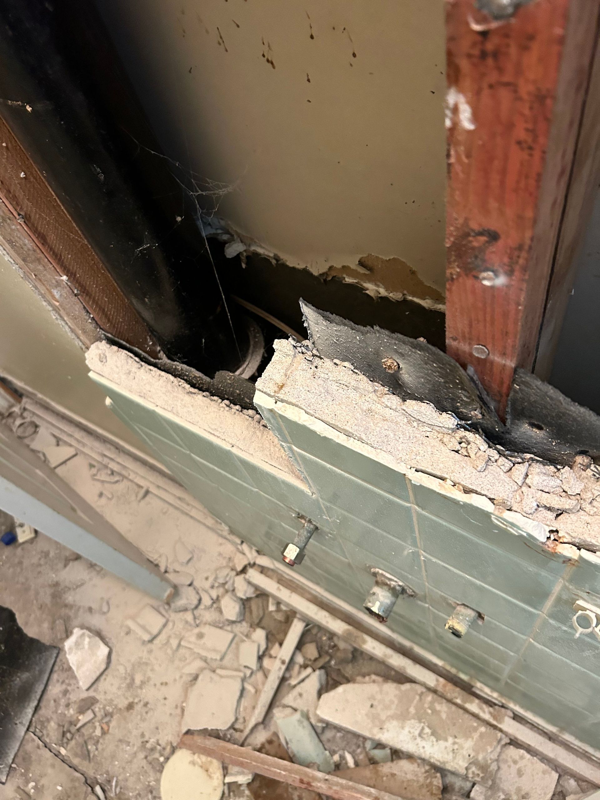 Damaged wall with exposed pipes and debris. Green tiles, brown wood, and beige ceiling are visible.