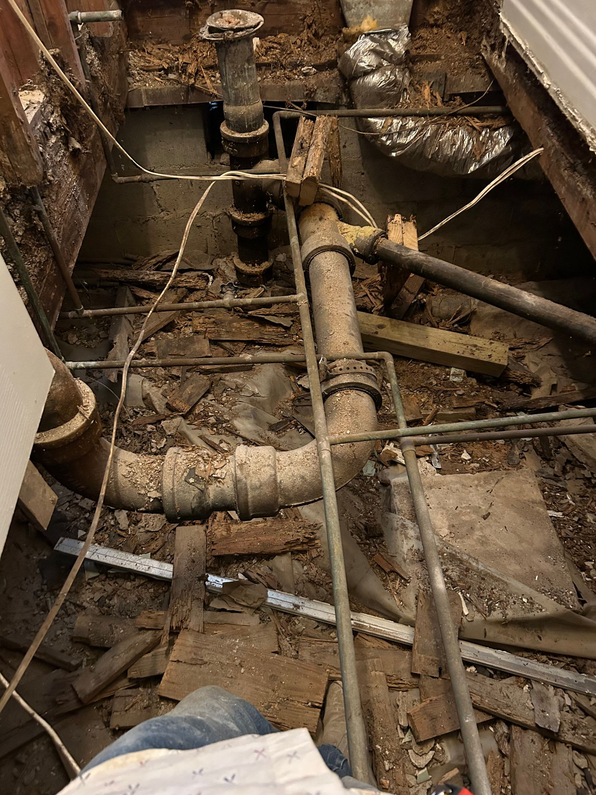 Plumbing and construction debris in an exposed crawlspace with pipes, wires, and wooden beams.