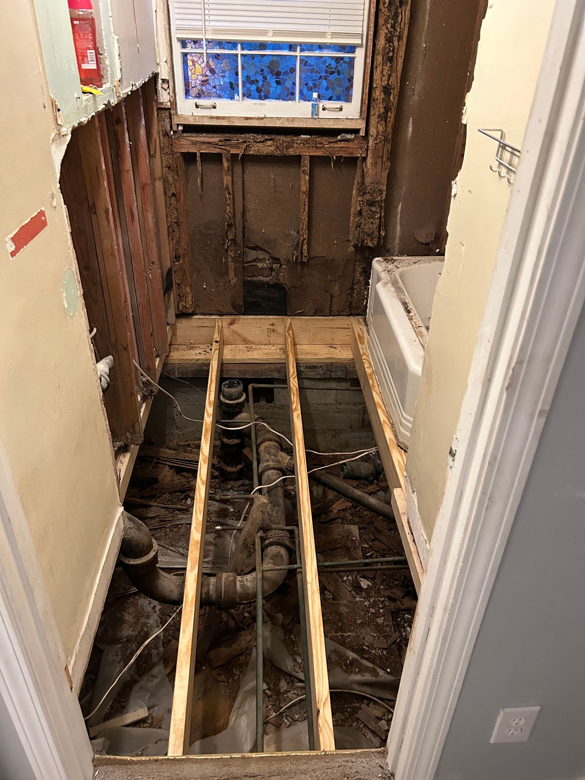 Bathroom renovation in progress; exposed pipes and floor joists; walls are bare, damaged.