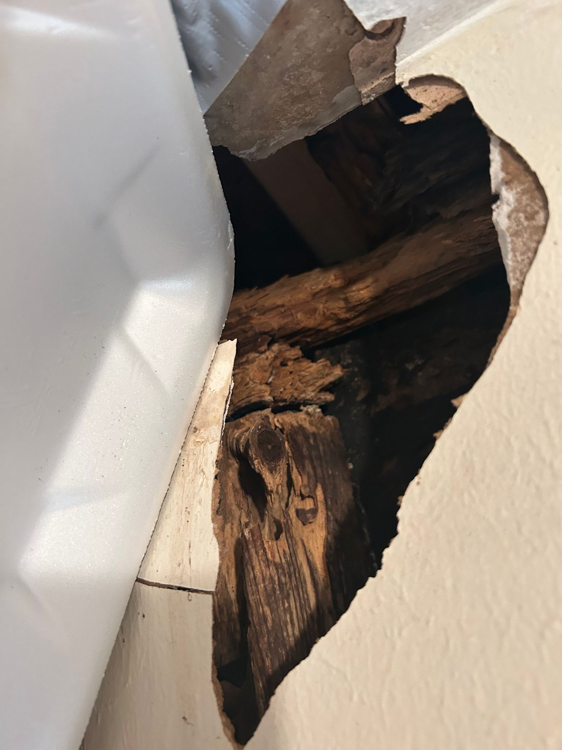Hole in wall, showing damaged, rotted wooden structure beneath. White wall to the right.