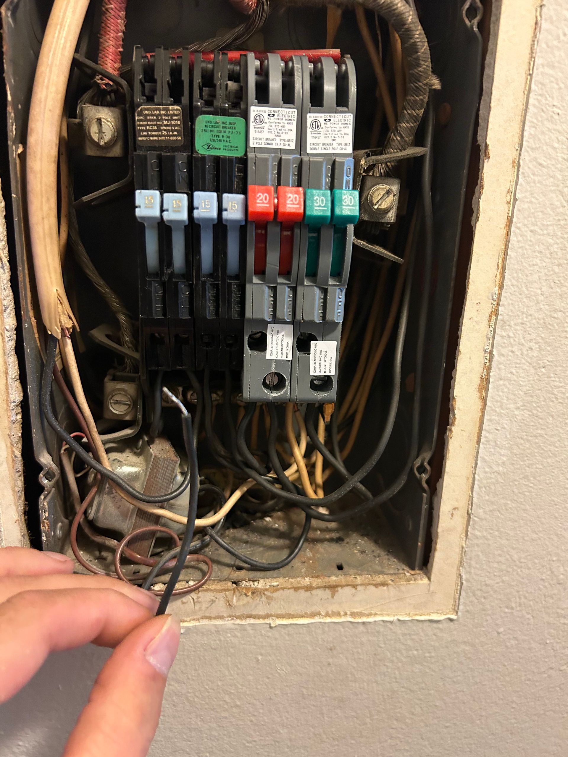 Hand holding a black wire near an old electrical panel with various colored fuses and wires in a wall.
