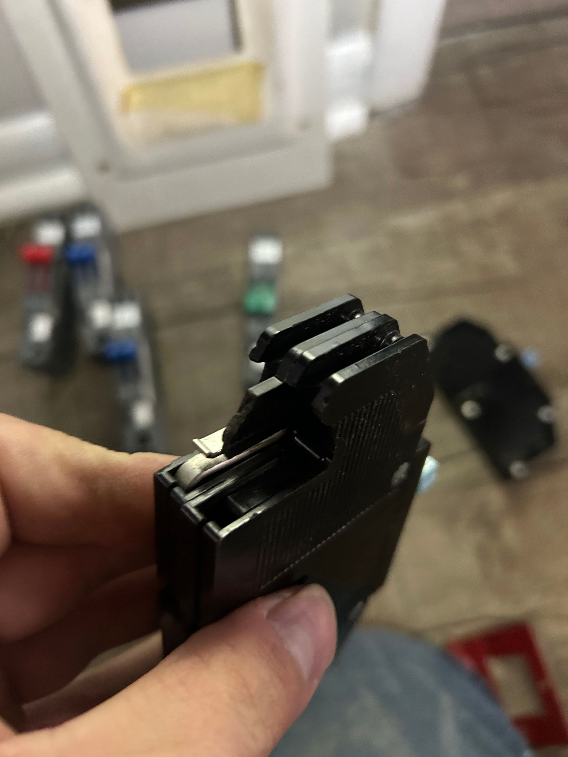 Hand holding a black plastic mechanism with a slotted top, against a blurred background of objects.