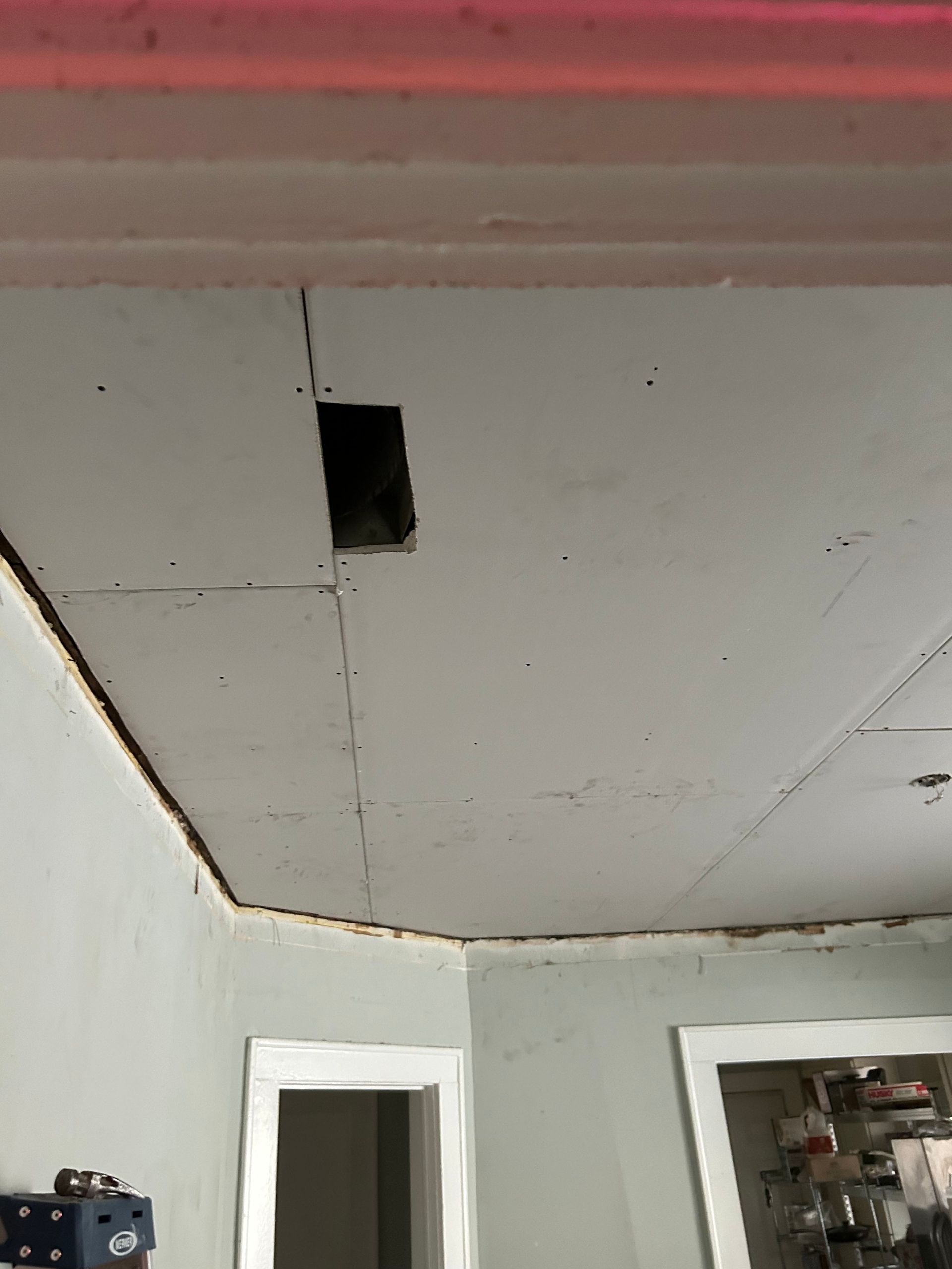 Ceiling with a hole, appears to be drywall, corner of a room, visible light fixture, and a door frame.