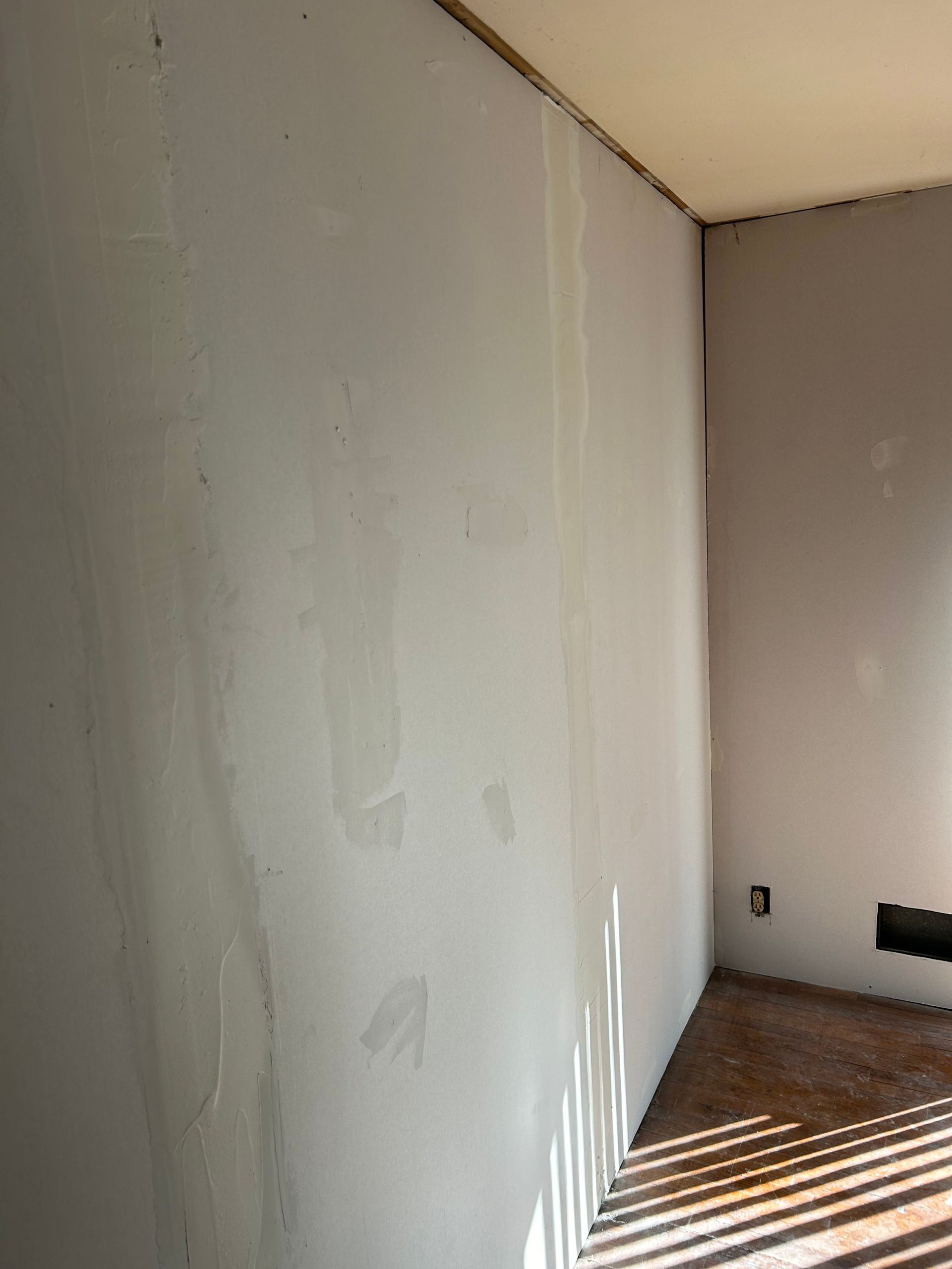 Interior view of a room with painted light gray walls. Light streams in, casting shadows on the floor.