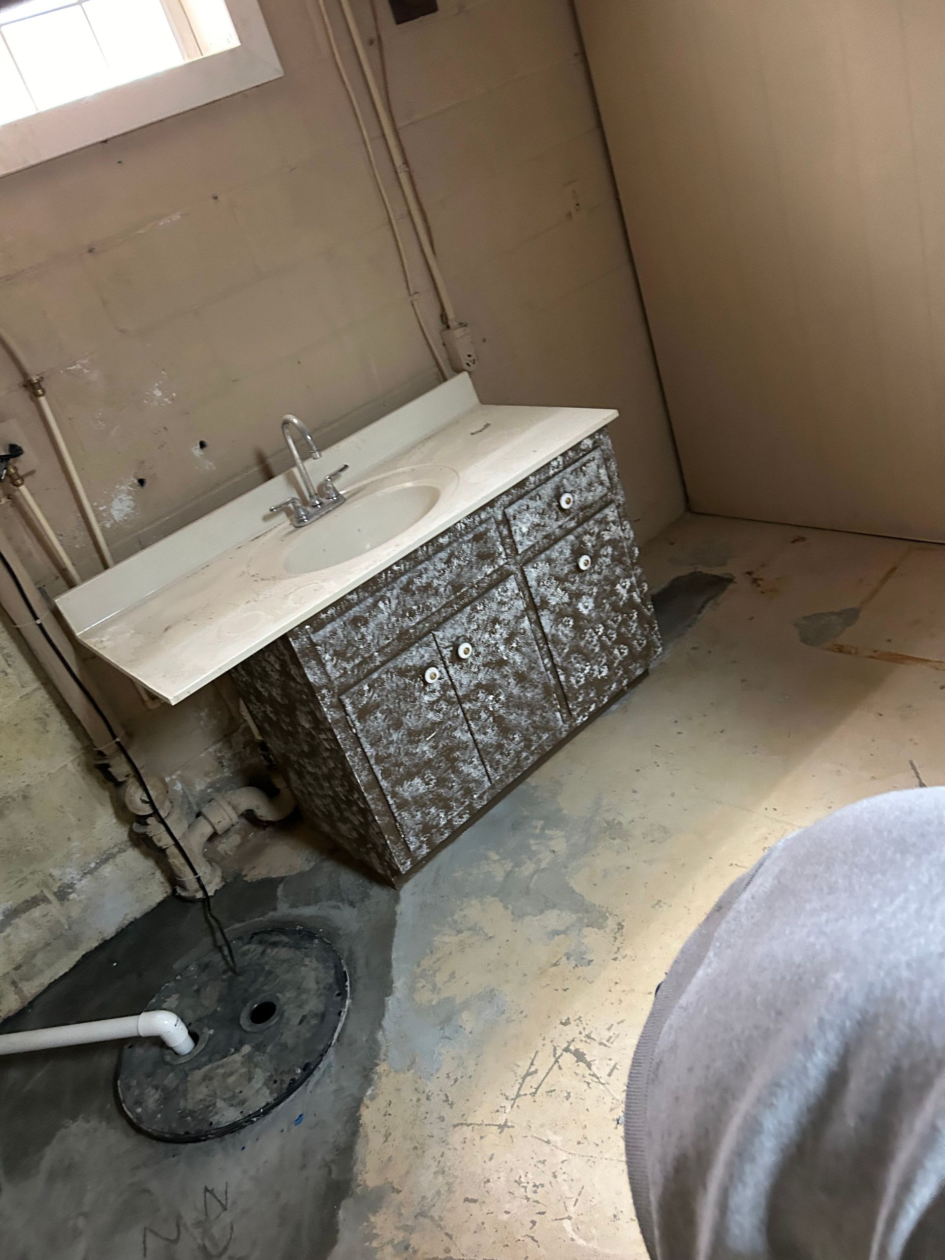 Basement room with a sink vanity, drain, and exposed pipes. Beige walls, gray flooring.
