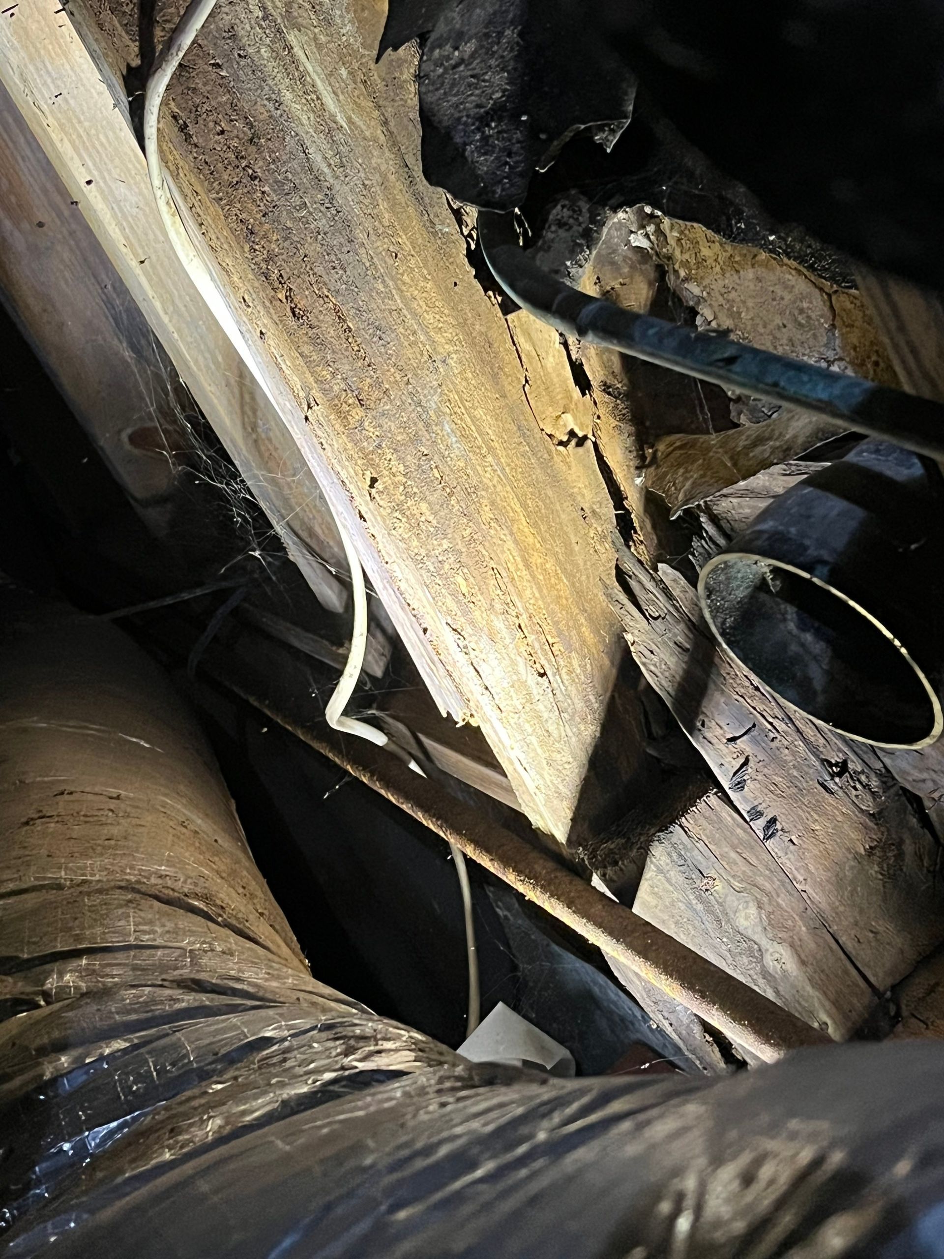 Dark, cluttered attic space. Wooden beams, pipes, and wiring are visible.