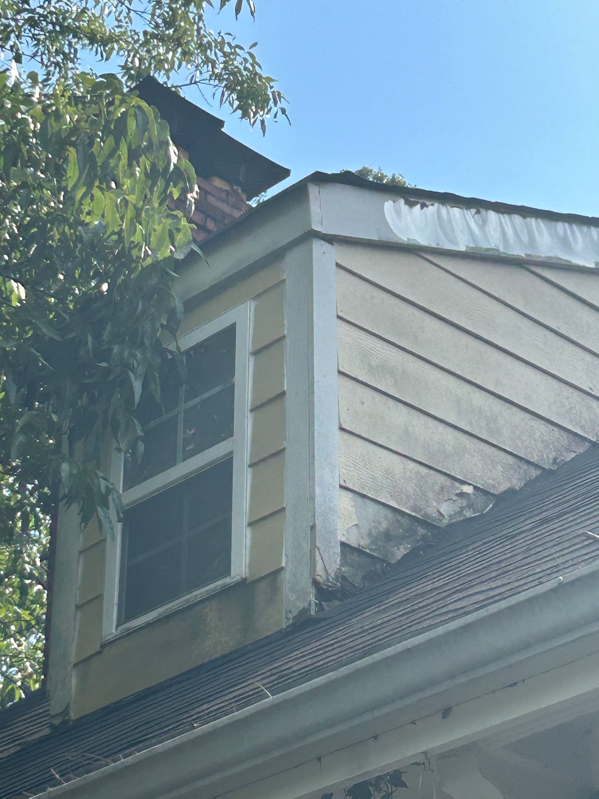 Dormer window with yellow siding and white trim, on a roof with dark shingles. A chimney sits above.