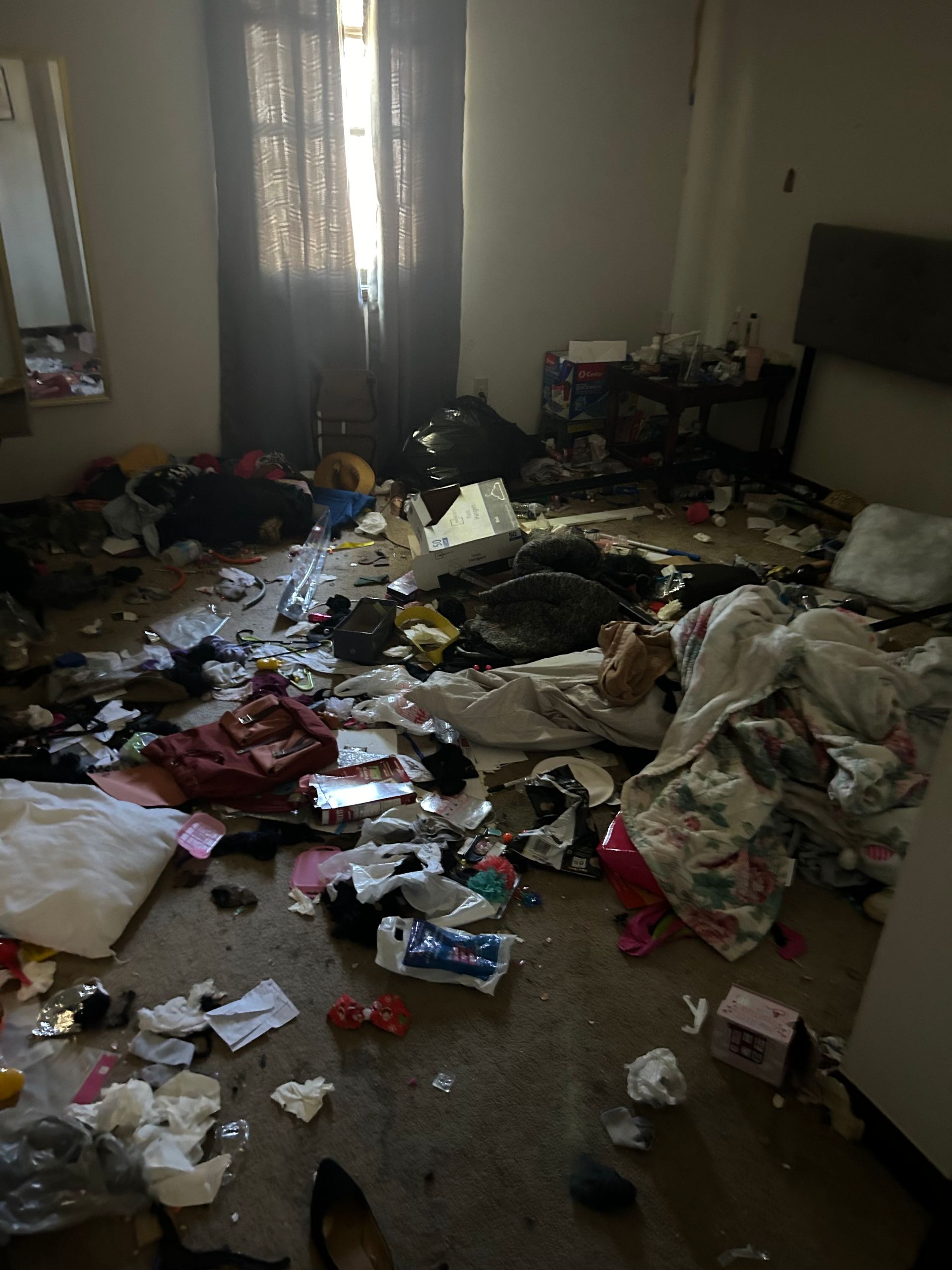 A cluttered bedroom. Clothing, trash, and other items are strewn across the floor. A window with light peeks through.
