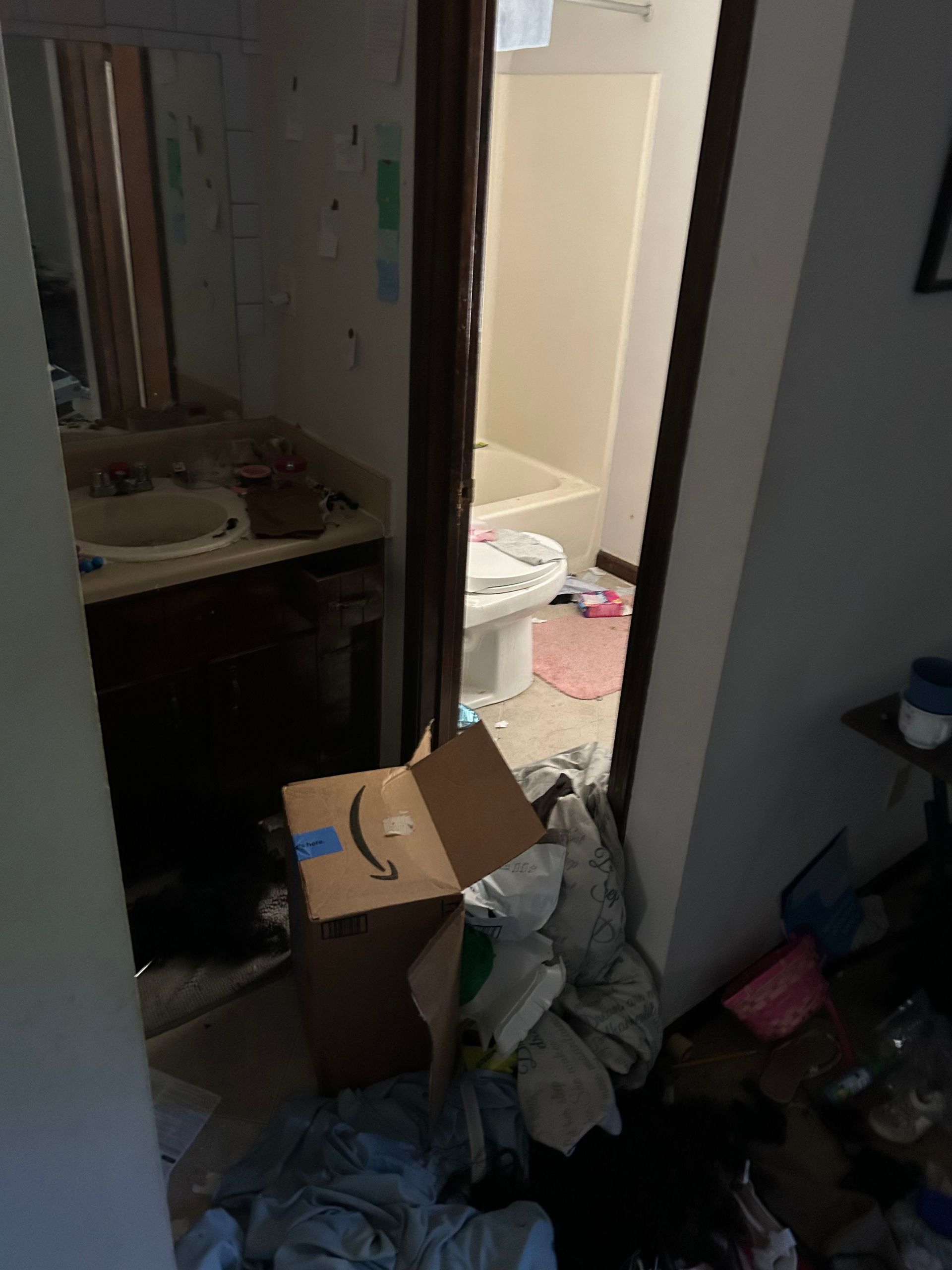 Messy bathroom: open doorway, cardboard box, clothing pile, toilet, and sink.