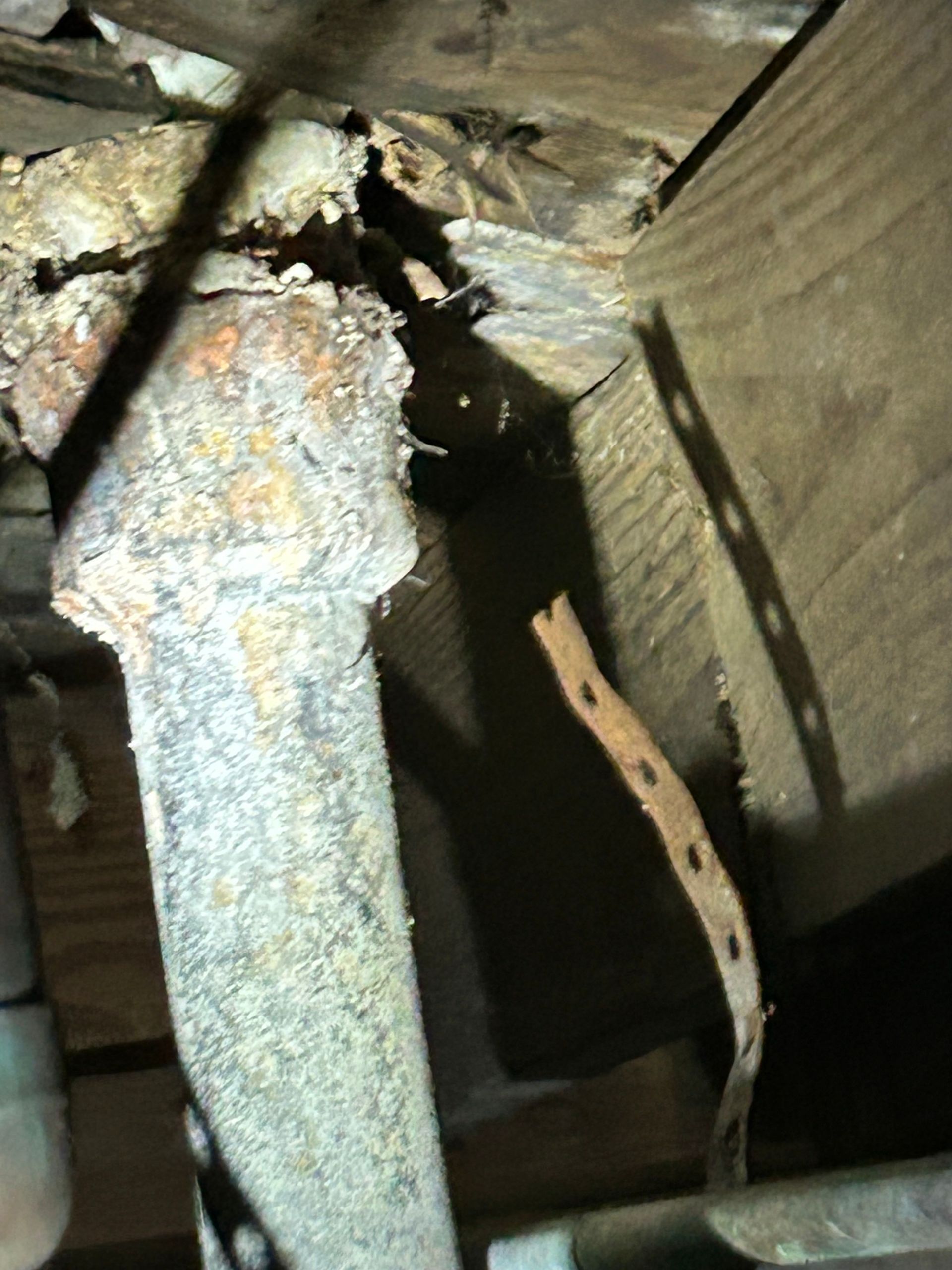 Rusty metal plumbing pipe in a dark, enclosed space, potentially a crawl space or under a floor.