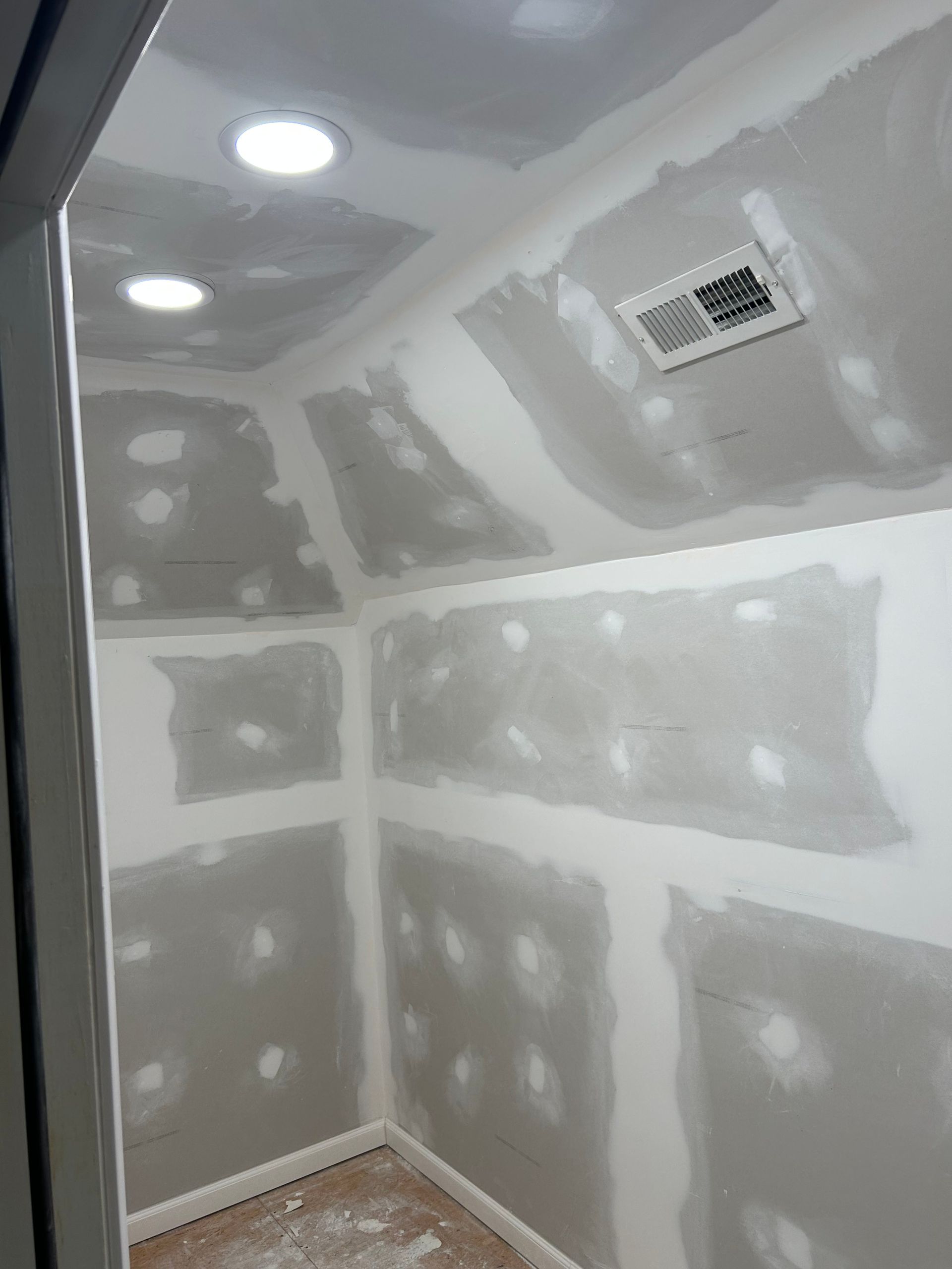 Interior of a small room under construction, with drywall installed. Two recessed lights and a vent are visible.