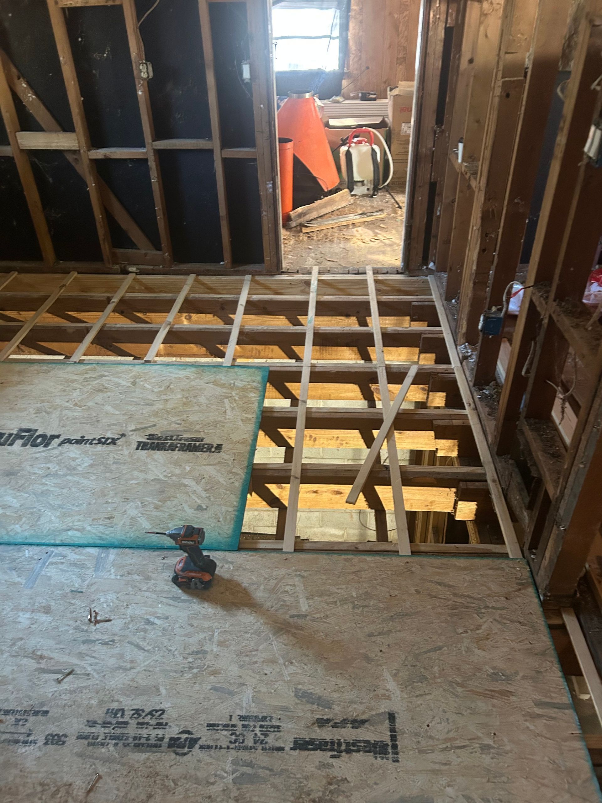 Interior view of a room under construction. Exposed floor joists, plywood, a power drill, and doorway in background.