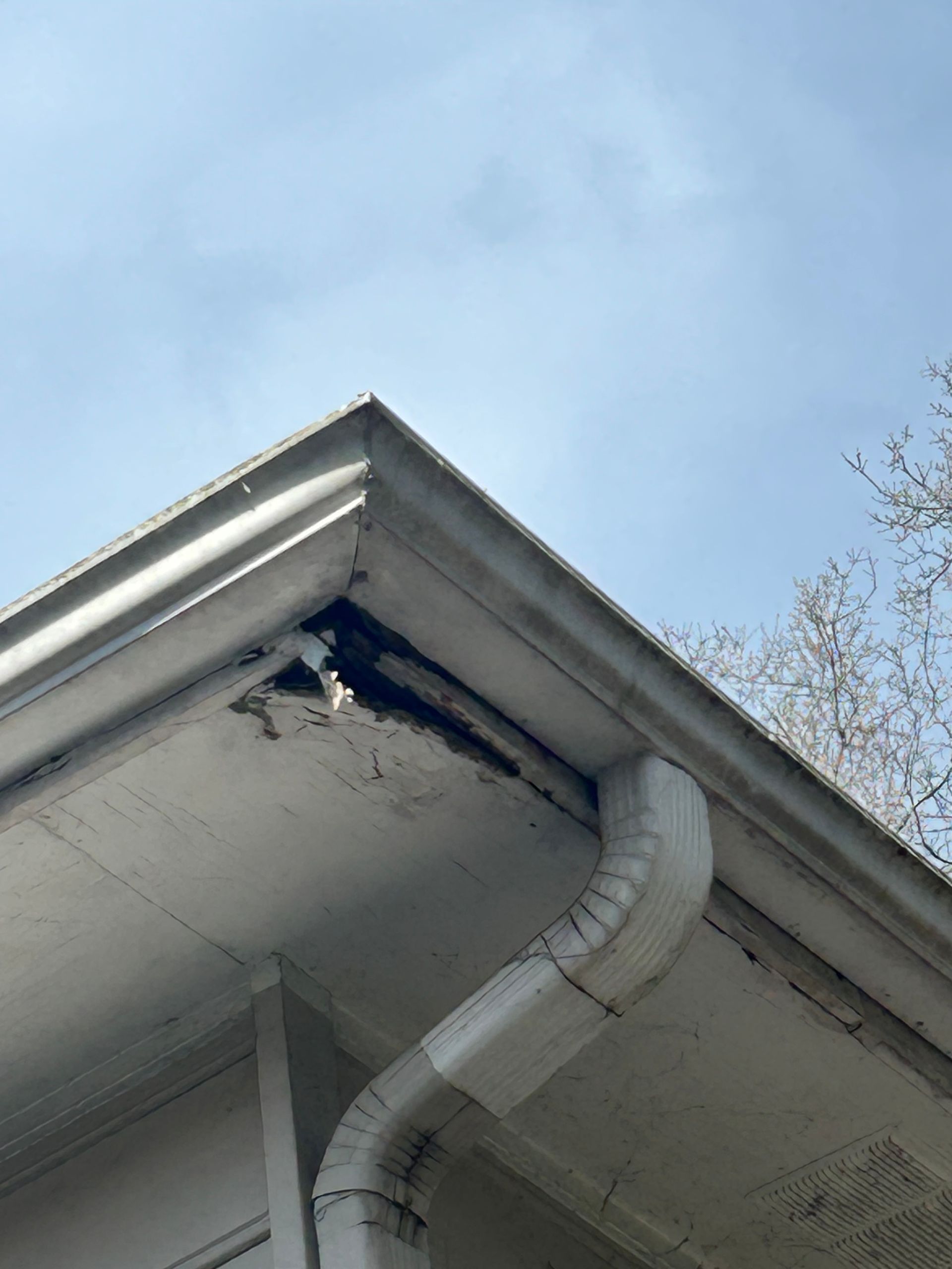 White gutter corner with damage, a downspout, and a cloudy sky in the background.