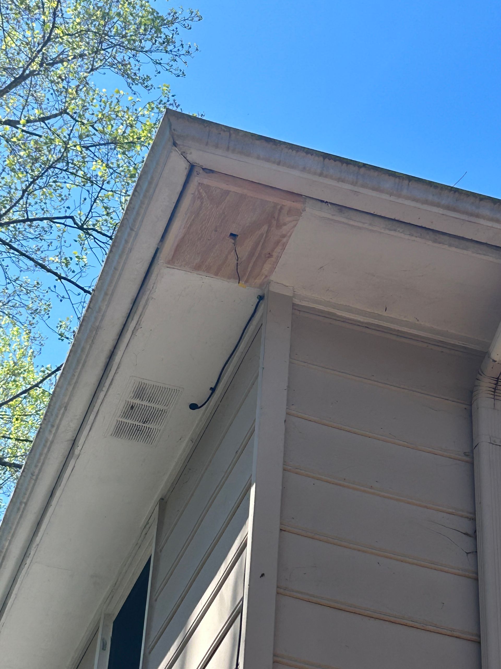 Exterior corner of a building with a damaged wooden soffit patch and a black cable, set against a blue sky.