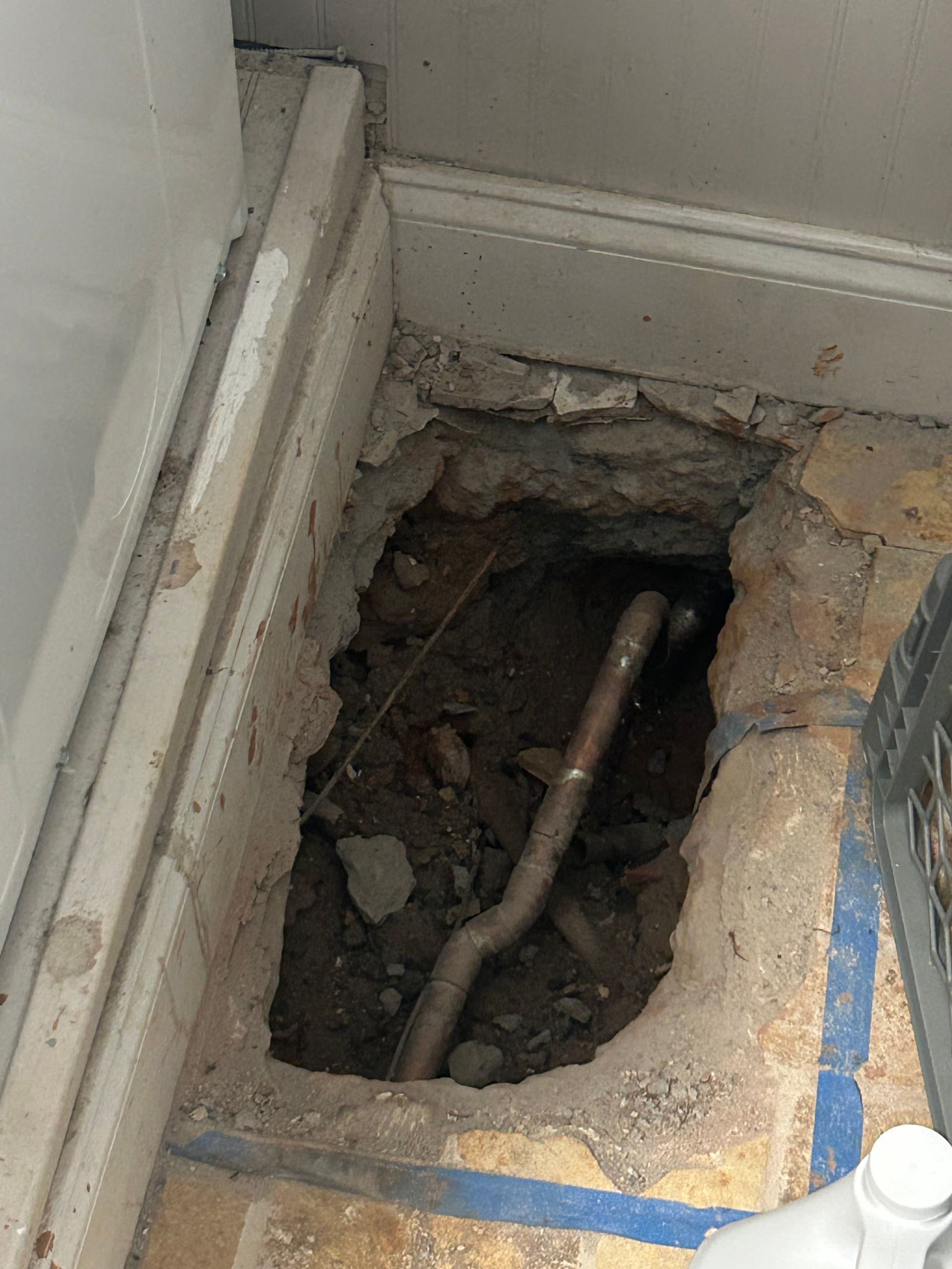 Hole in floor revealing pipes; concrete floor with blue line, dirt, and plumbing visible.