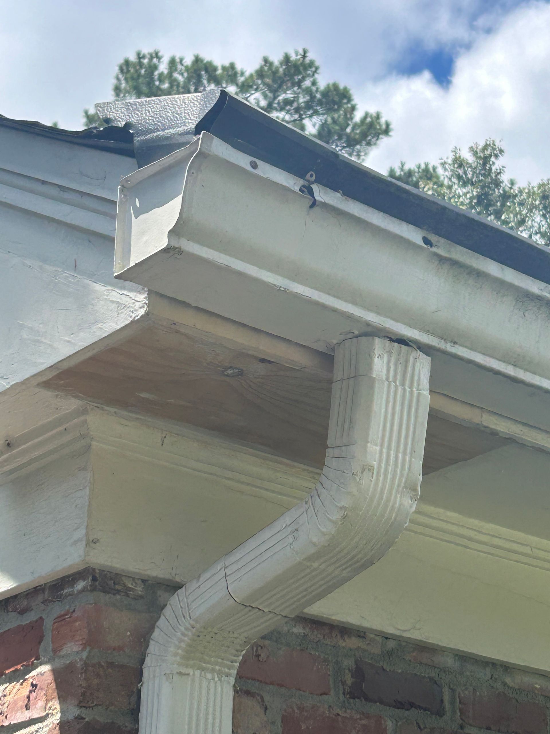 White gutter and downspout attached to a brick building; a section of wood paneling sits beneath the gutter.