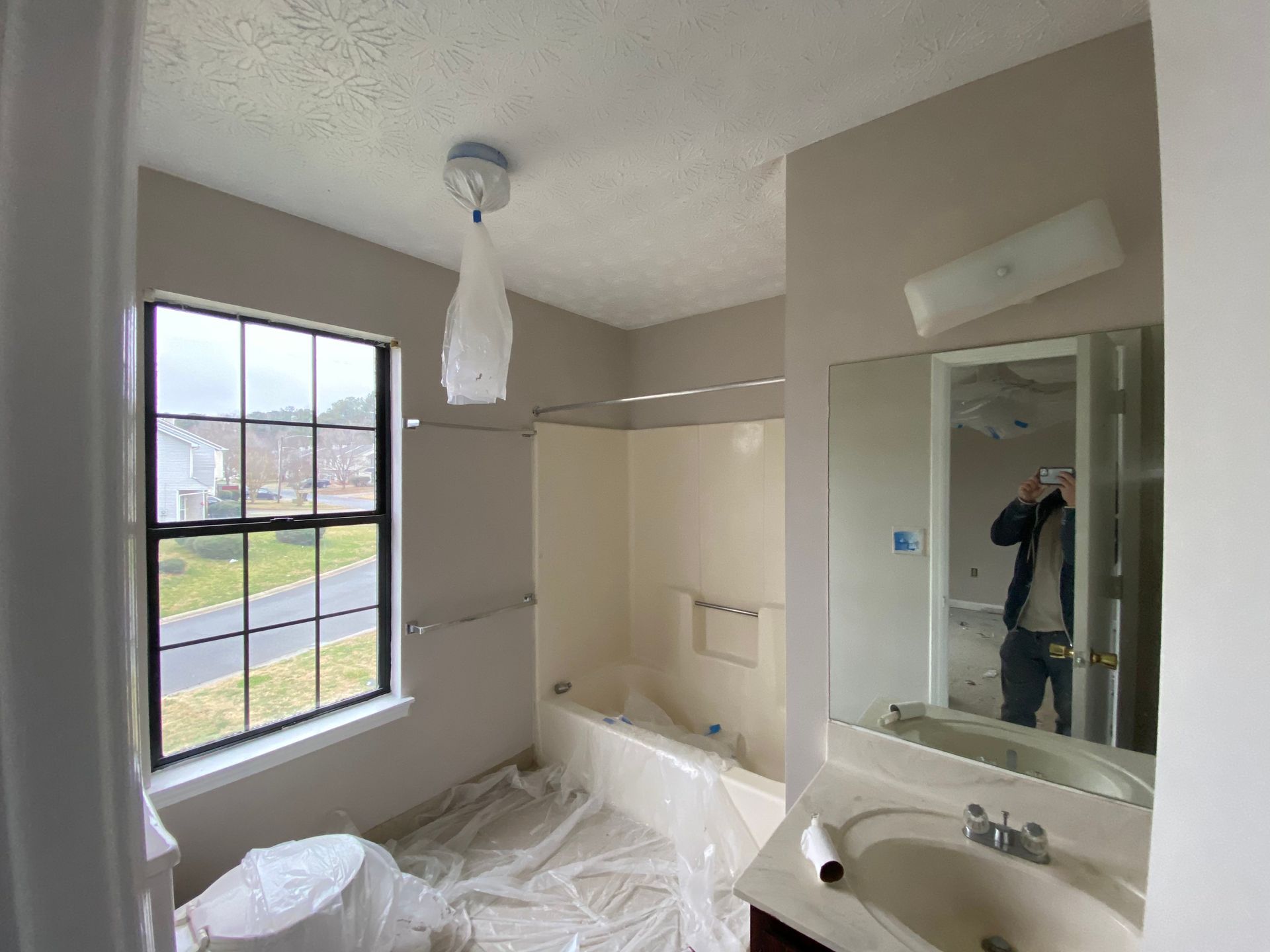 Bathroom under renovation, beige walls, shower and toilet covered in plastic, window, person in mirror.