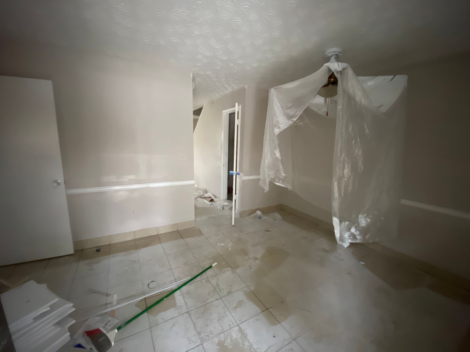 Room under renovation; light-colored walls, tile floor, plastic sheeting, door open to hallway, mop leaning against a wall.