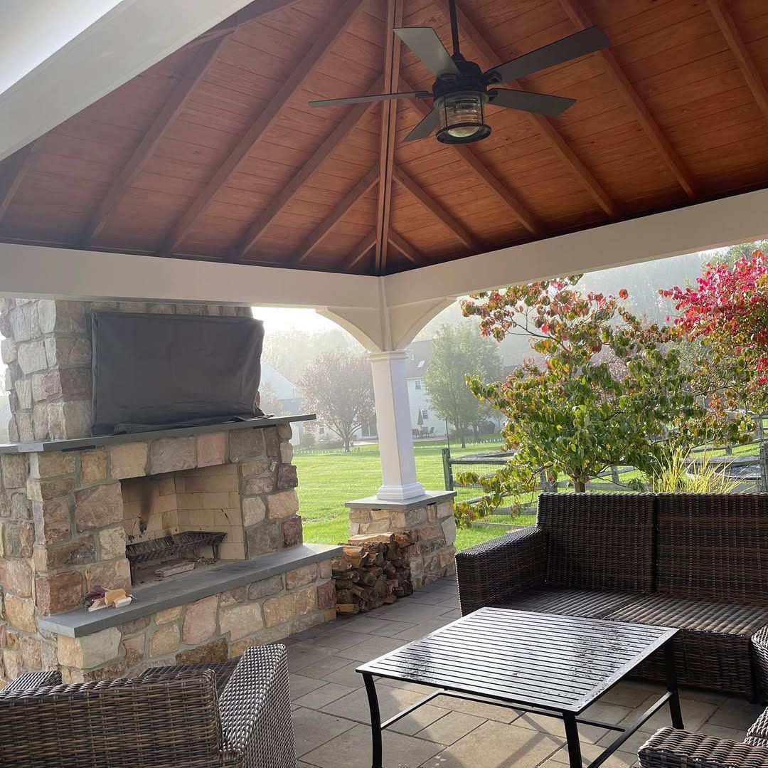 A gazebo with a fireplace , couch , table and ceiling fan.