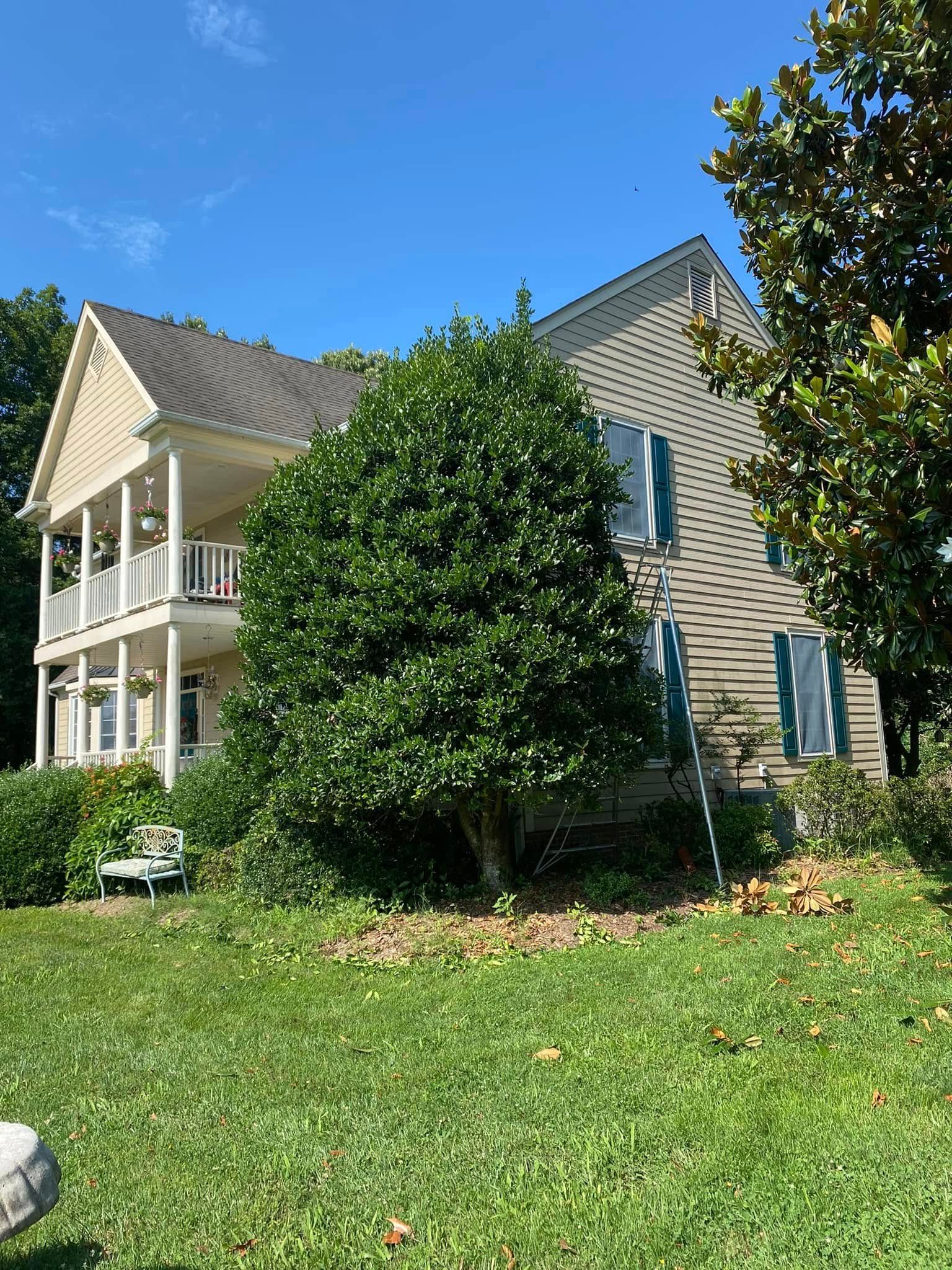 A two-story, light-colored house with a wrap-around porch, surrounded by lush green trees and a lawn on a sunny day.