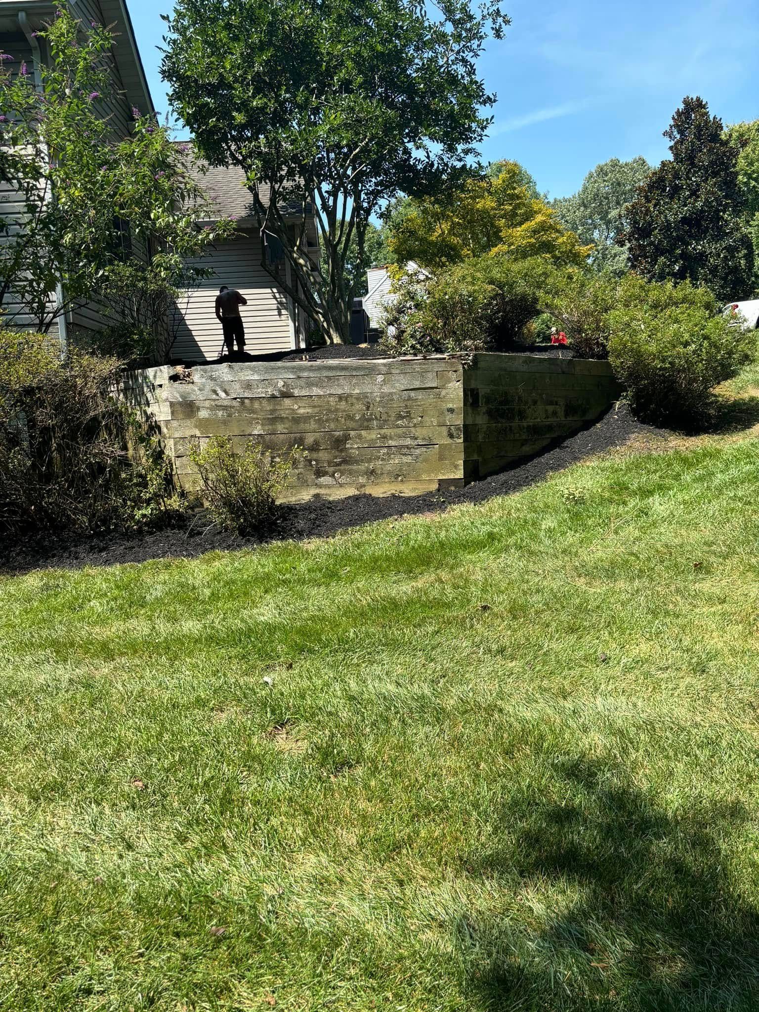 A retaining wall made of stacked stone blocks sits in a sunny backyard next to a residential house and landscaping.