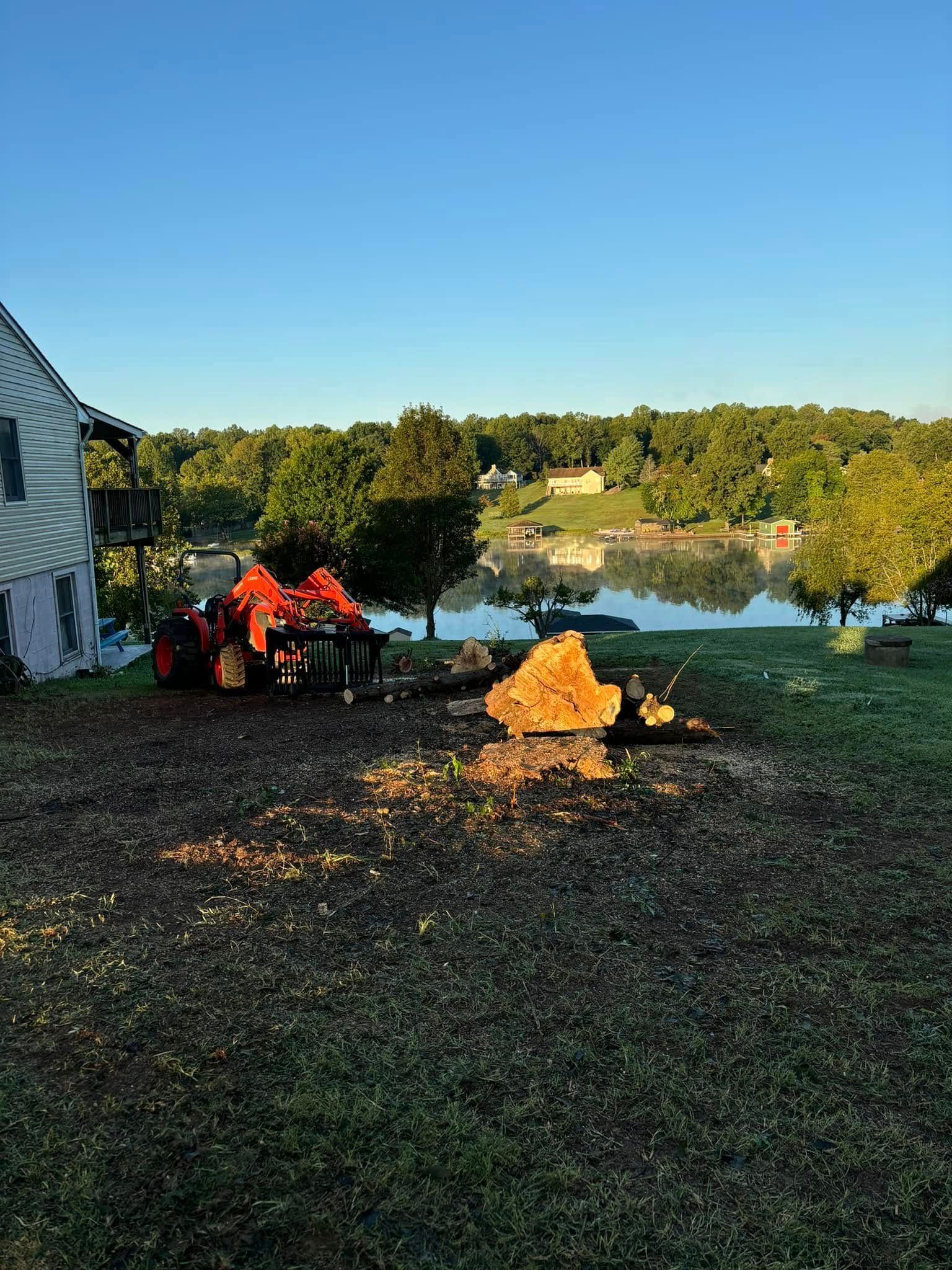 An orange tractor sits on a grassy lawn near a tree stump, with a house on the left and a lake in the background.