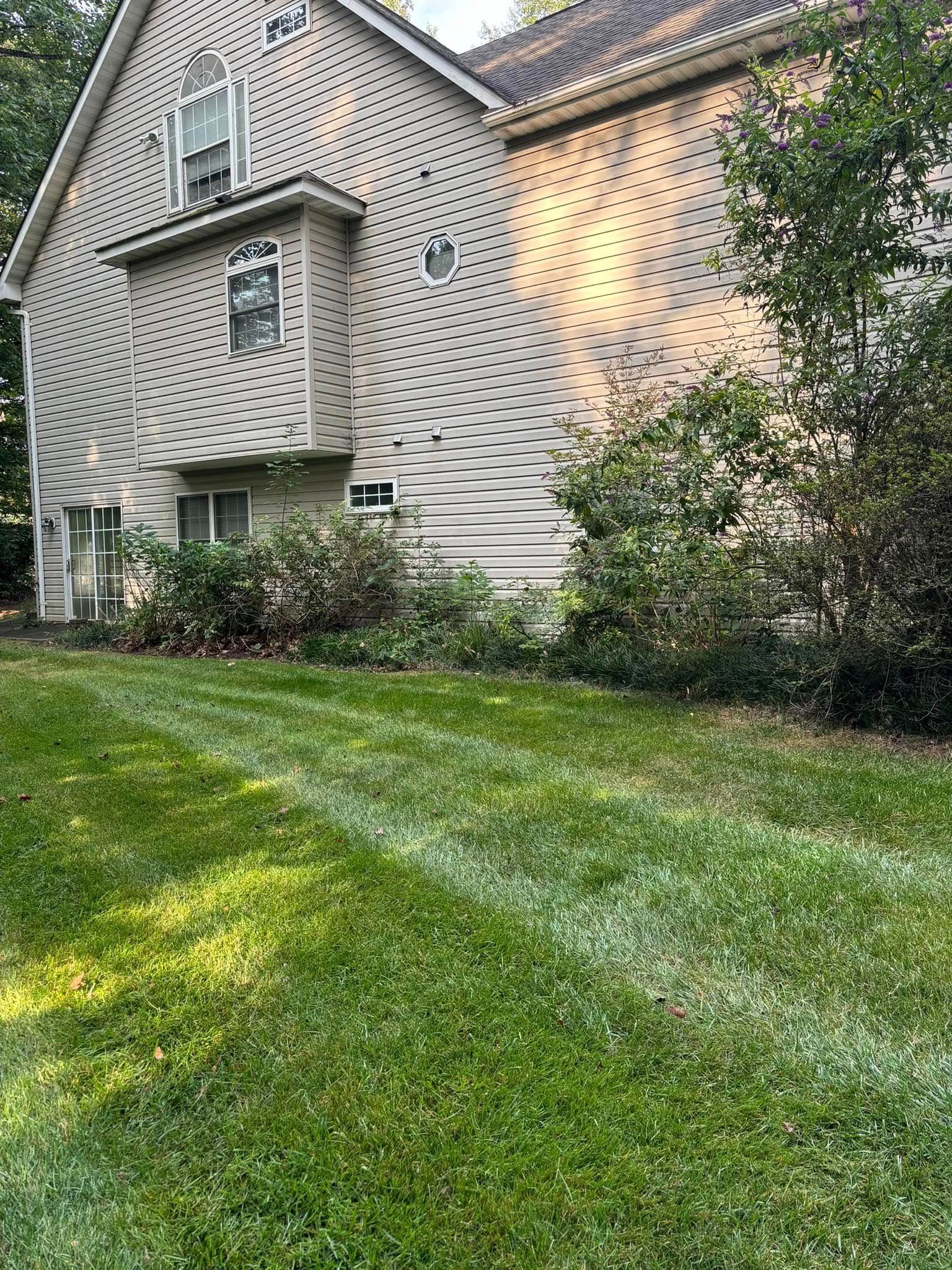 Side view of a two-story beige house with a bay window, bordered by a green lawn and shrubs under dappled sunlight.