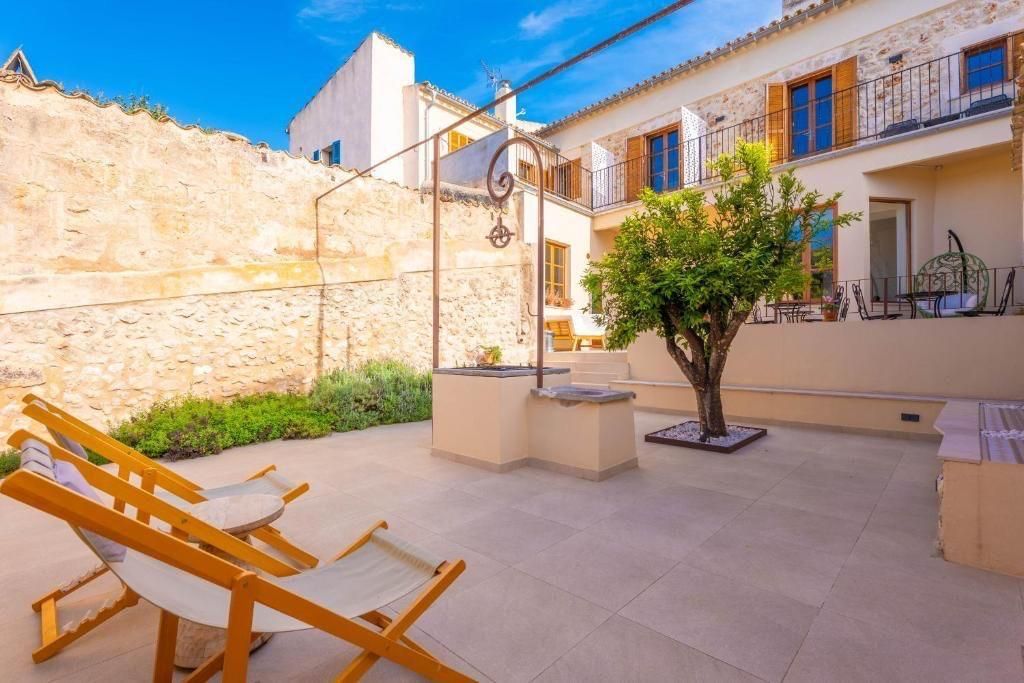 Dos tumbonas de madera en un patio con un pozo, un árbol y un edificio al fondo.