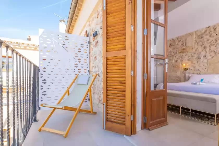 Balcón con tumbona, contraventanas de madera y vistas a un dormitorio con cama.