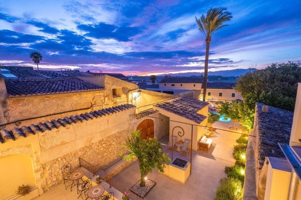Patio iluminado al atardecer, con edificios de piedra, palmera y piscina. Cielo púrpura y naranja.