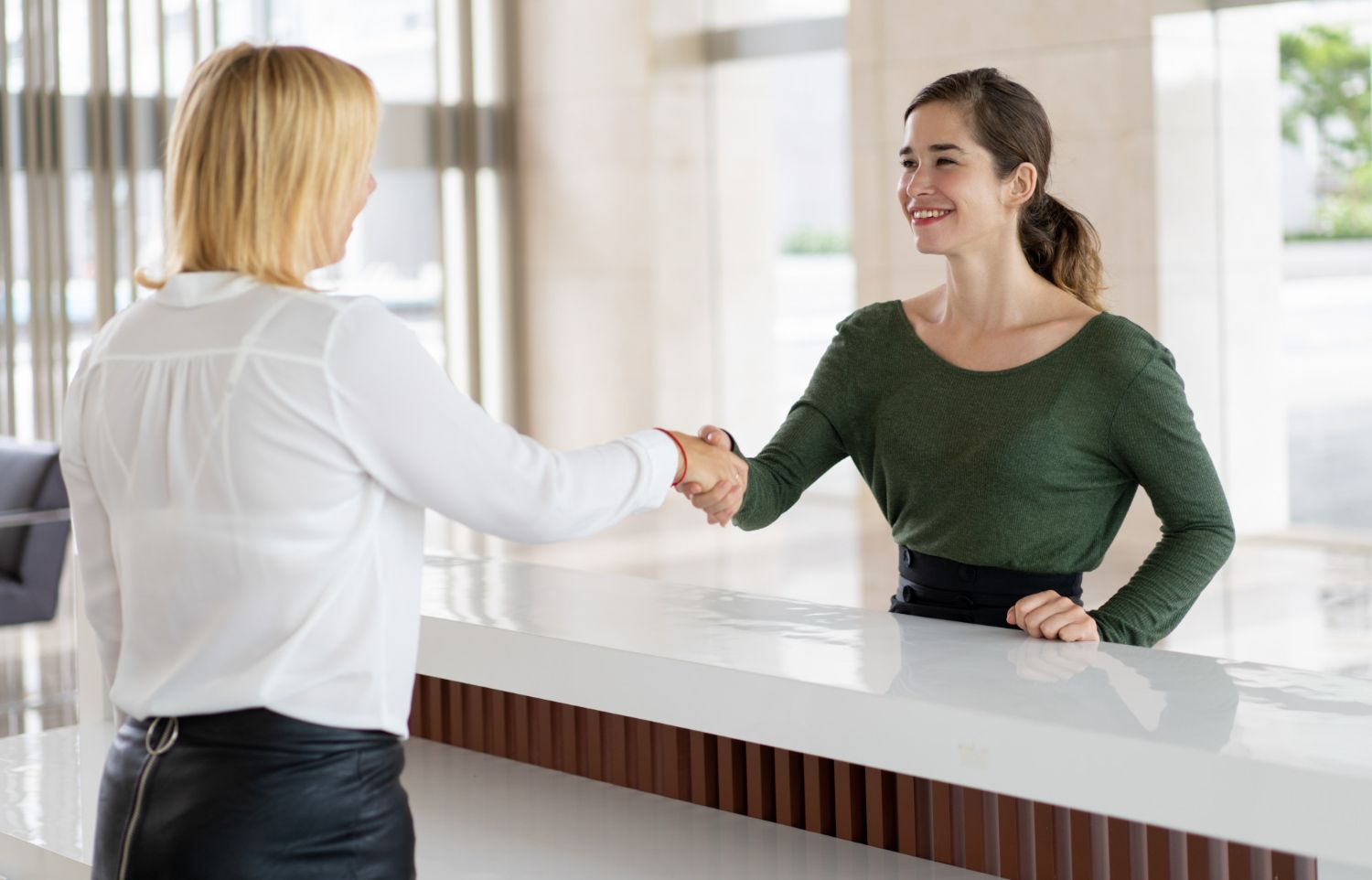 Woman shaking hands with receptionist at a white counter; interior, smiles.