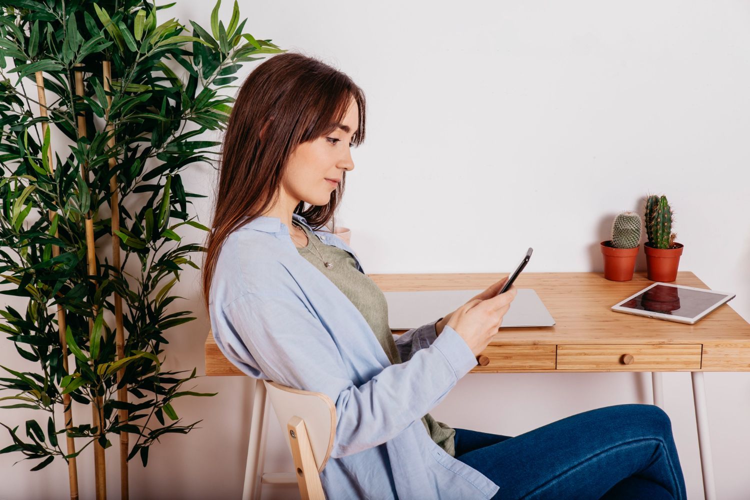 Woman sits at a desk, looking at a tablet. Plants and laptop are on the desk.