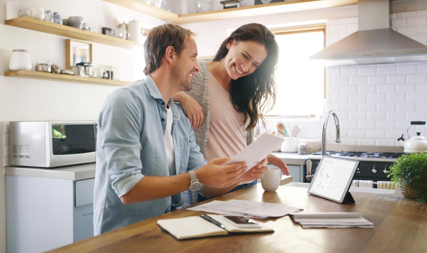 Couple in kitchen reviewing documents, smiling. Man is holding papers, woman leans over him, light-filled setting.