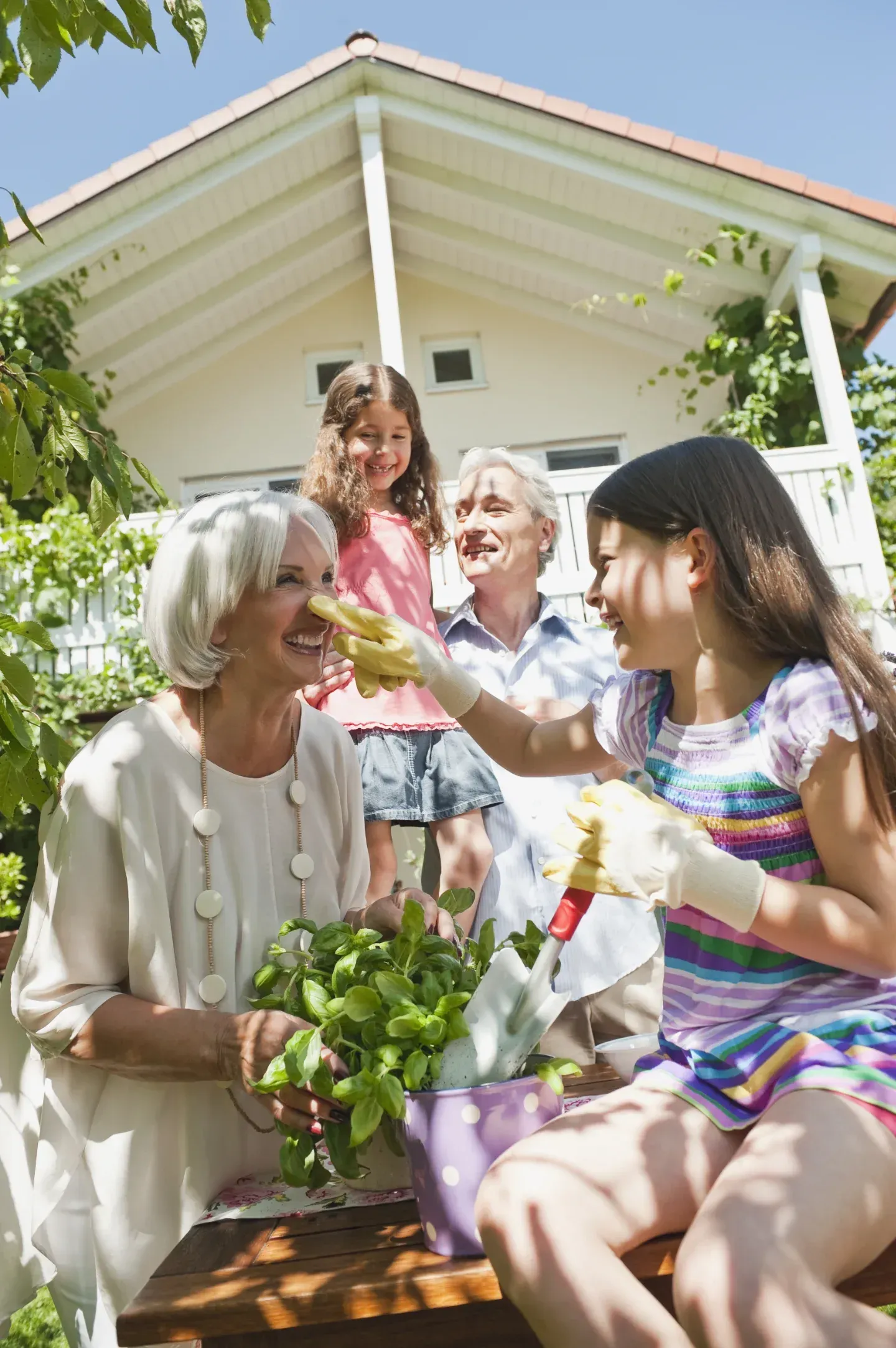 Family gardening together outdoors; child touches grandmother's face with plant.