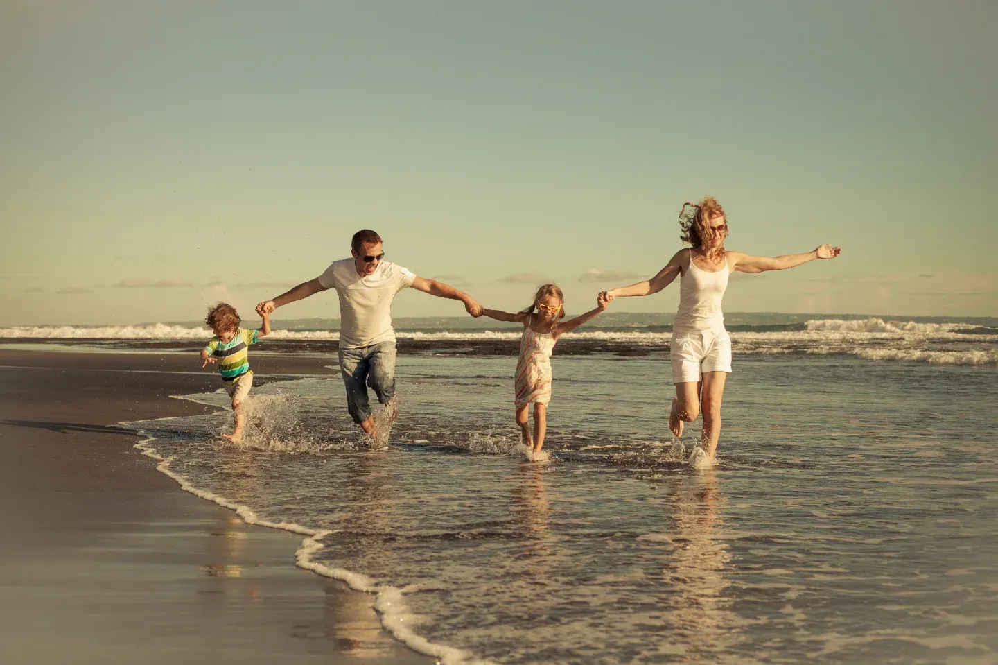 Family of four running on a beach, holding hands with arms outstretched; sunny day.